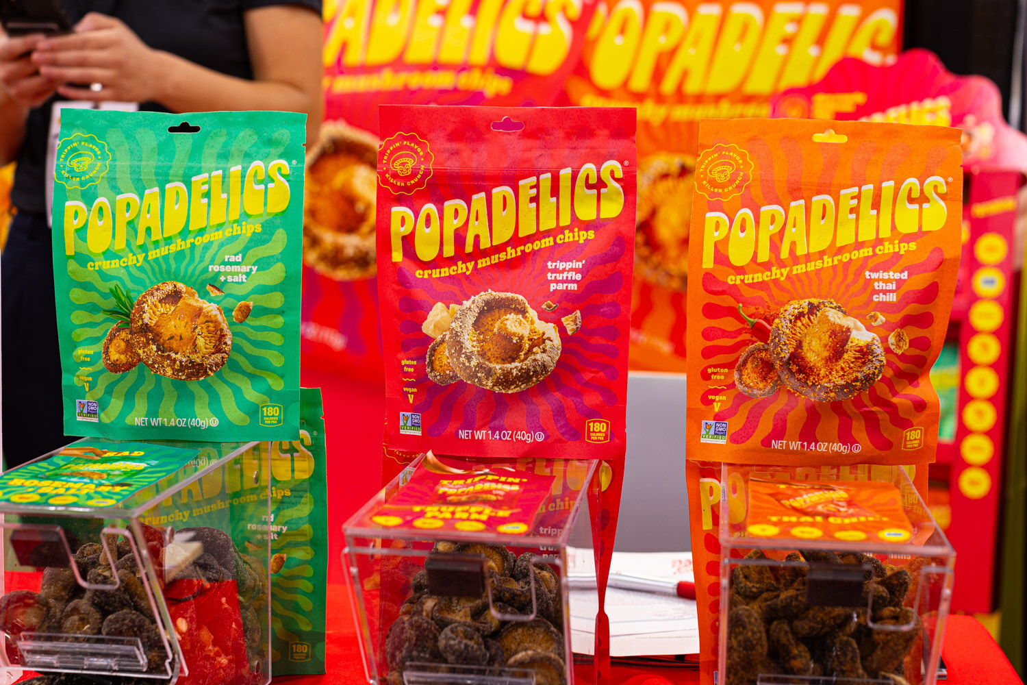Colorful Popadelics crunchy mushroom chip bags stand above clear bins of snacks on a red and yellow display