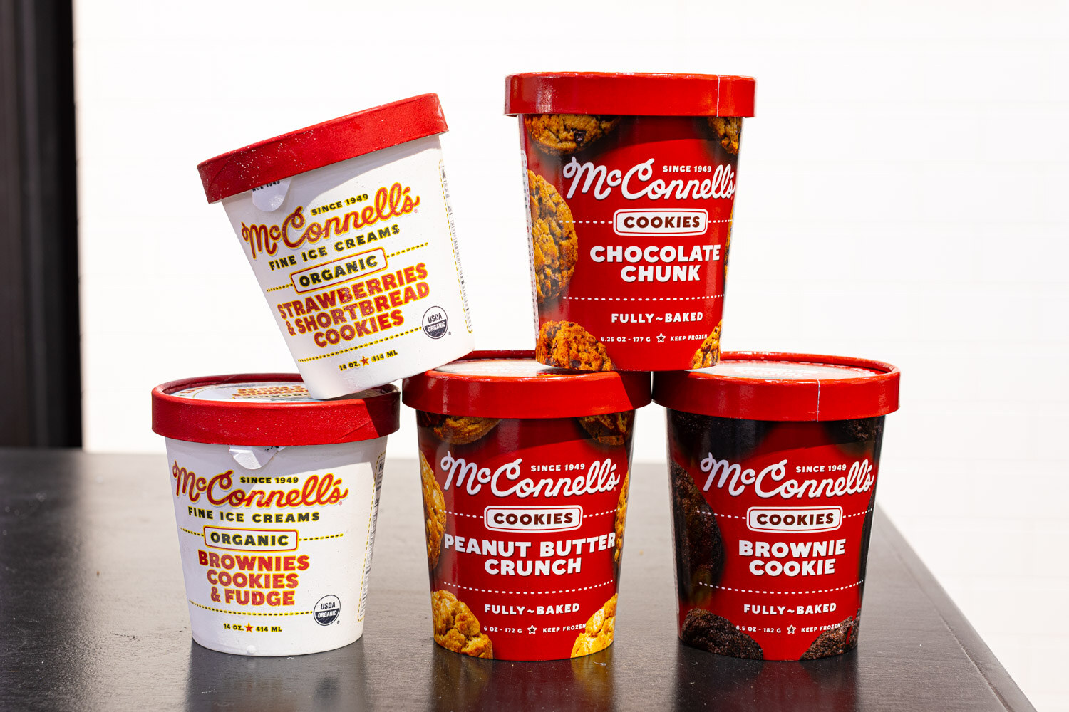 Stacked pints of McConnell's Fine Ice Creams cookie flavors, including Chocolate Chunk and Brownie Cookie, on a dark table