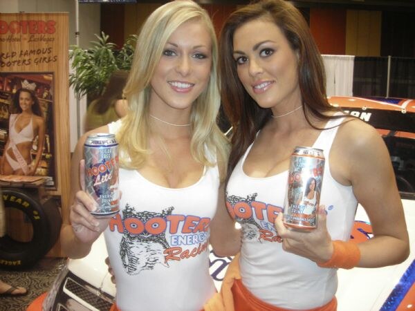 Two smiling women, one blonde and one brunette, wear white Hooters Energy Racing tops and each hold a can of Hooters energy drink, one marked Lite. A race car and Hooters banner are behind them.