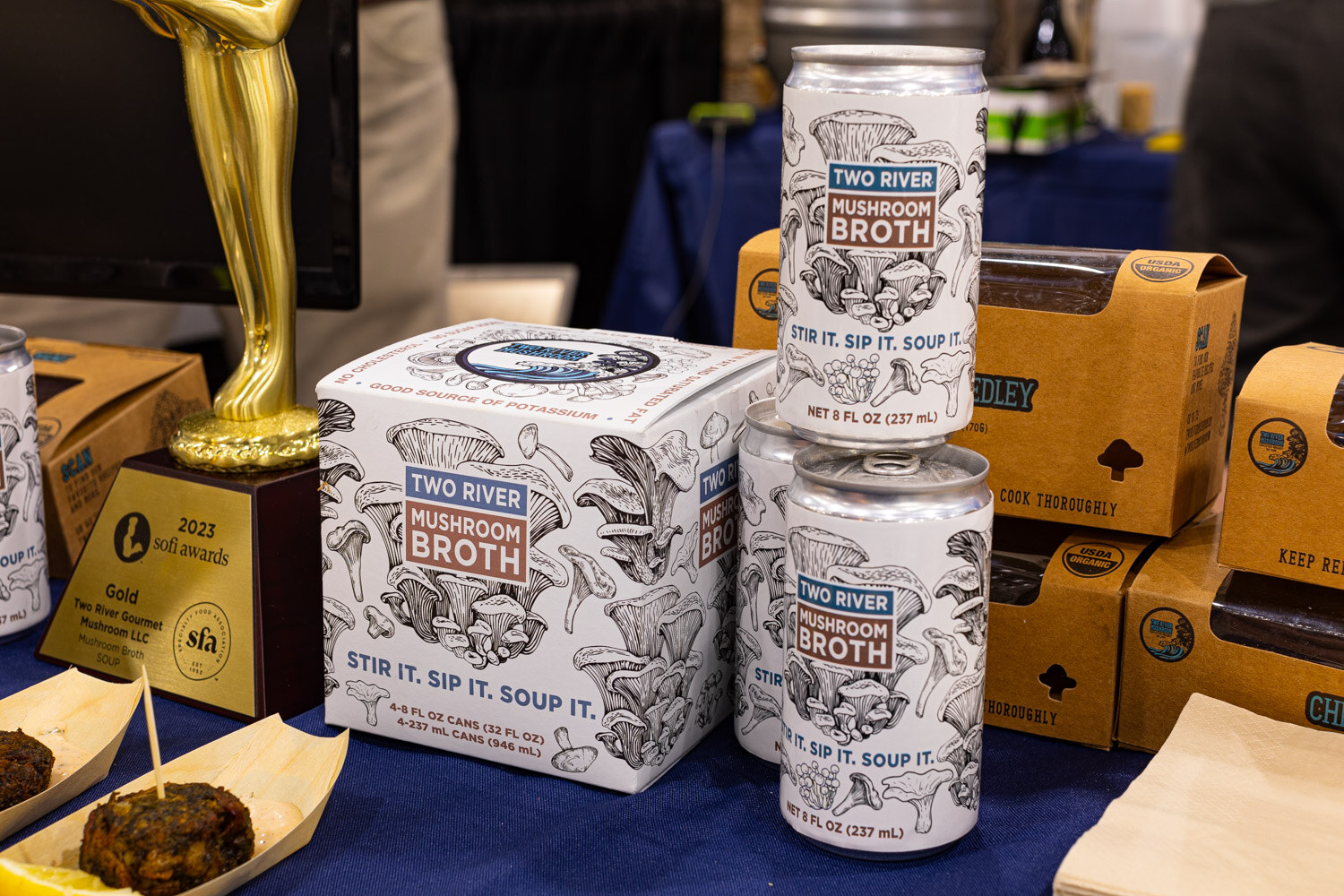 Cans and boxes of Two River Mushroom Broth sit beside a gold award trophy on a navy tablecloth