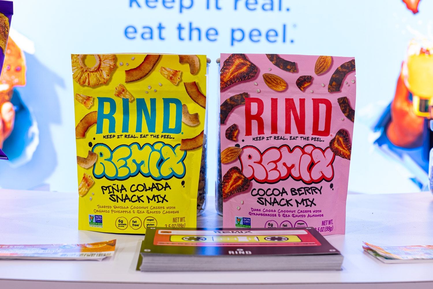 Two RIND Remix bags in Pina Colada and Cocoa Berry flavors stand in front of a sign reading keep it real, eat the peel