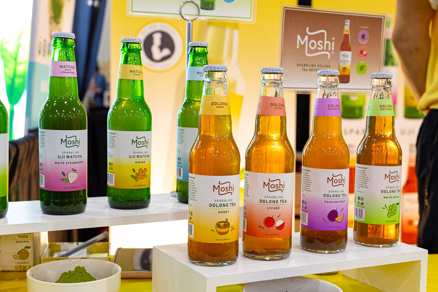 Bottles of Moshi sparkling tea and oolong tea in assorted flavors stand on a white shelf beside a Moshi sign