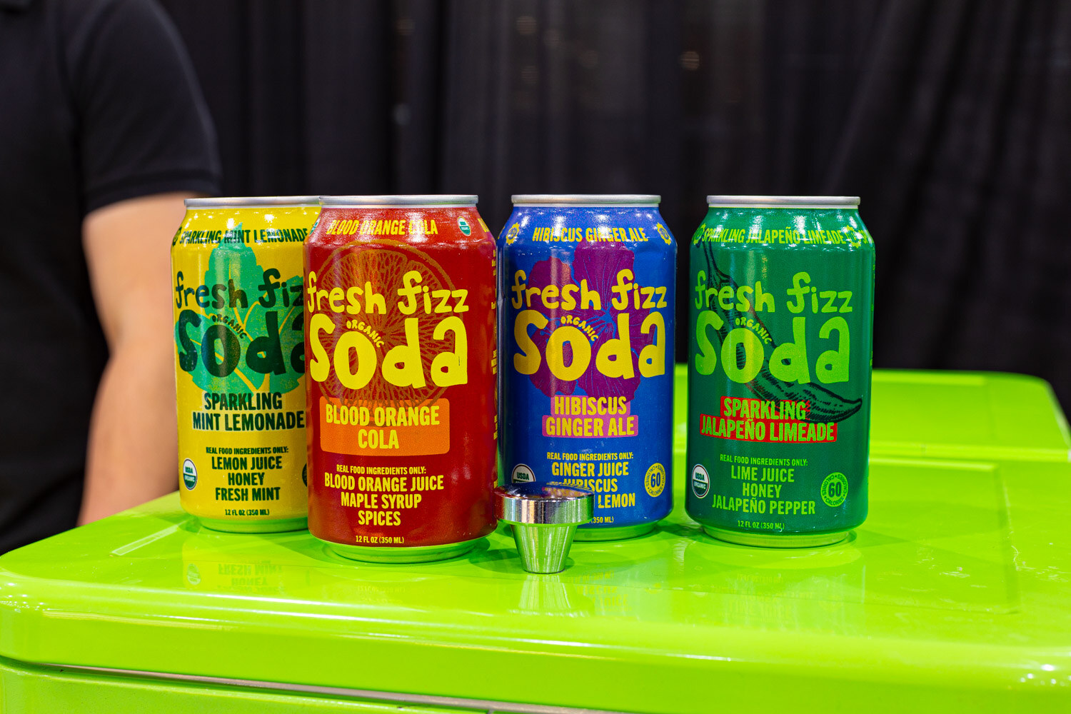 Cans of Fresh Fizz Soda in Mint Lemonade, Blood Orange Cola, Hibiscus Ginger Ale and Lime Jalapeno flavors sit on a lime green cooler