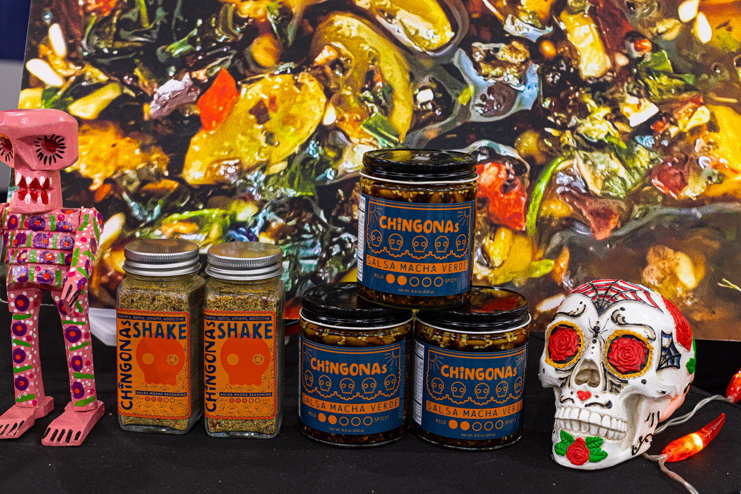 Jars of Chingona Shake seasoning sit beside colorful skull figurines in front of a vivid illustrated backdrop