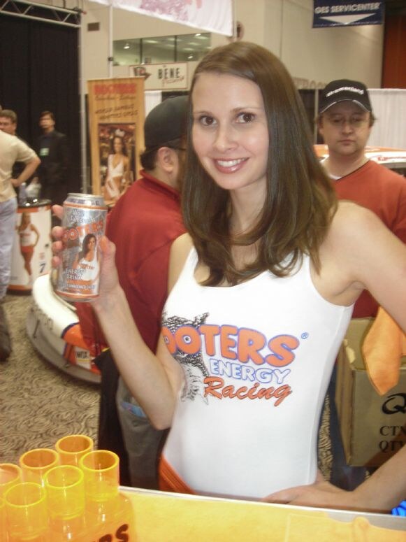 A smiling woman with long brown hair holds up a Hooters energy drink can and wears a white Hooters Energy Racing top. Orange cups sit on the counter, with other attendees and a race car behind her.