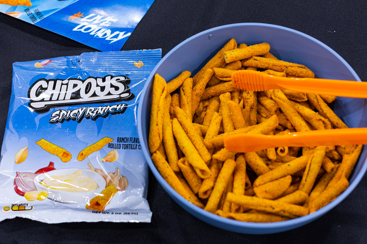 A bag of Chipoys Spicy Ranch next to a blue bowl of orange rolled tortilla chips with tongs