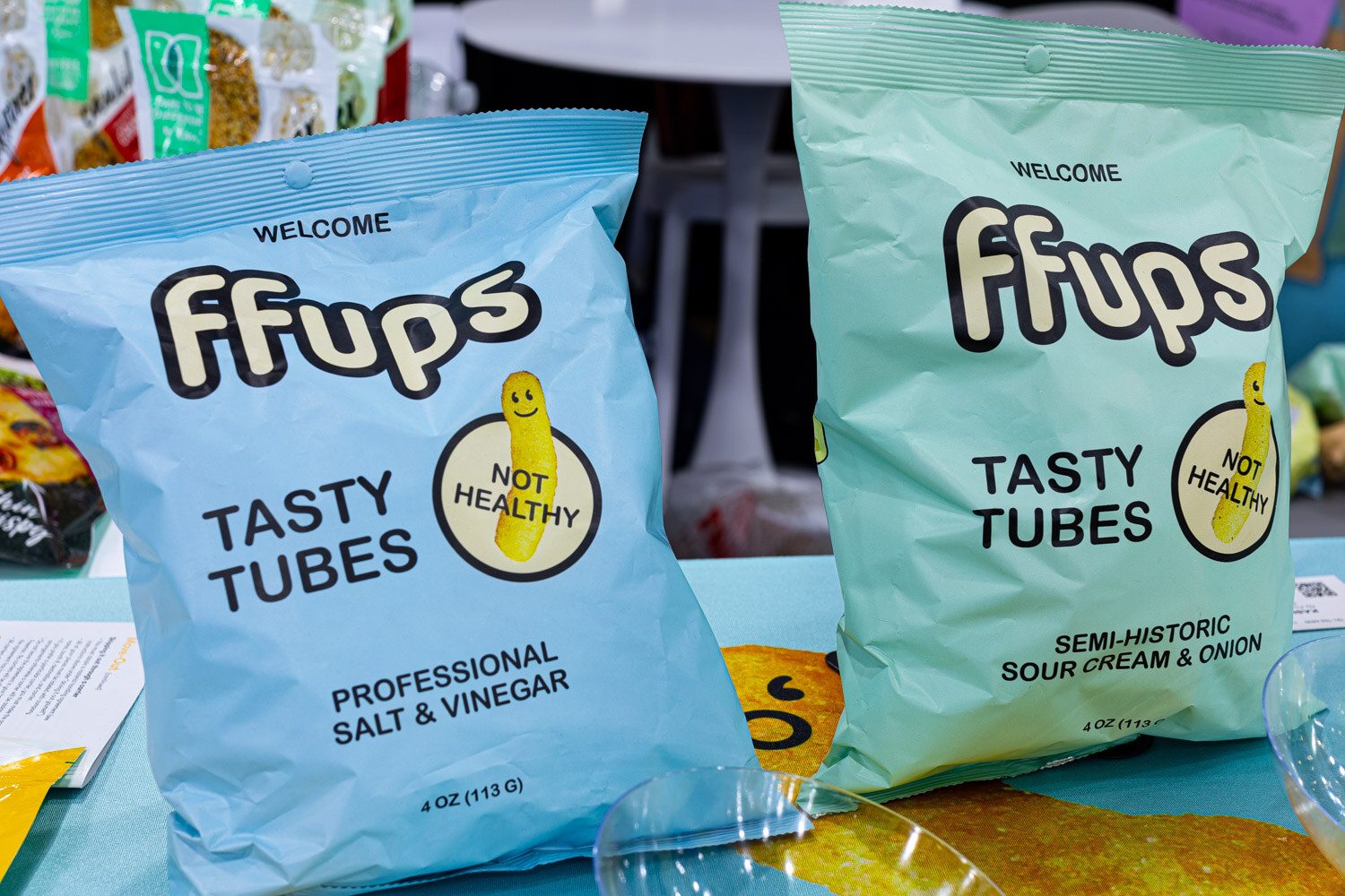 Two blue ffups Tasty Tubes bags in Salt and Vinegar and Sour Cream and Onion flavors, labeled Not Healthy, sit on a teal table
