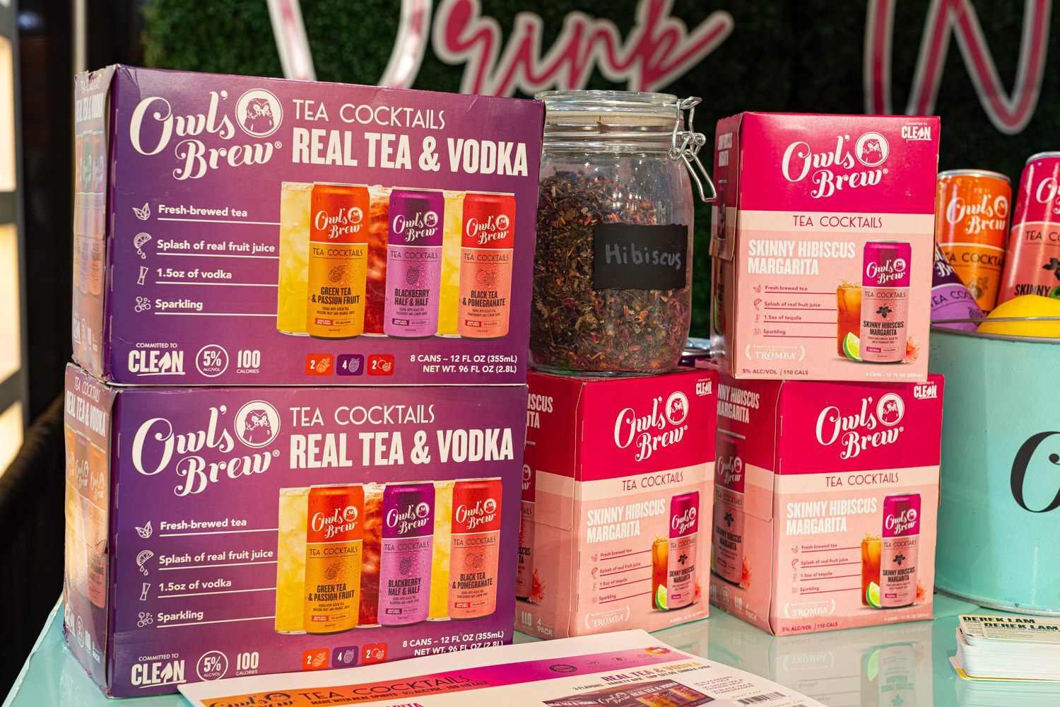 Stacked boxes of Owl's Brew Real Tea and Vodka tea cocktails in purple and pink packaging beside a glass jar of loose tea