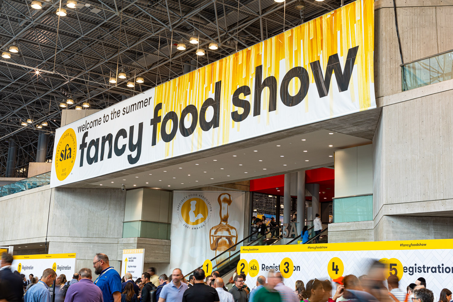 A large yellow and white welcome to the summer fancy food show banner hangs over the entrance at the Javits Center as attendees queue below