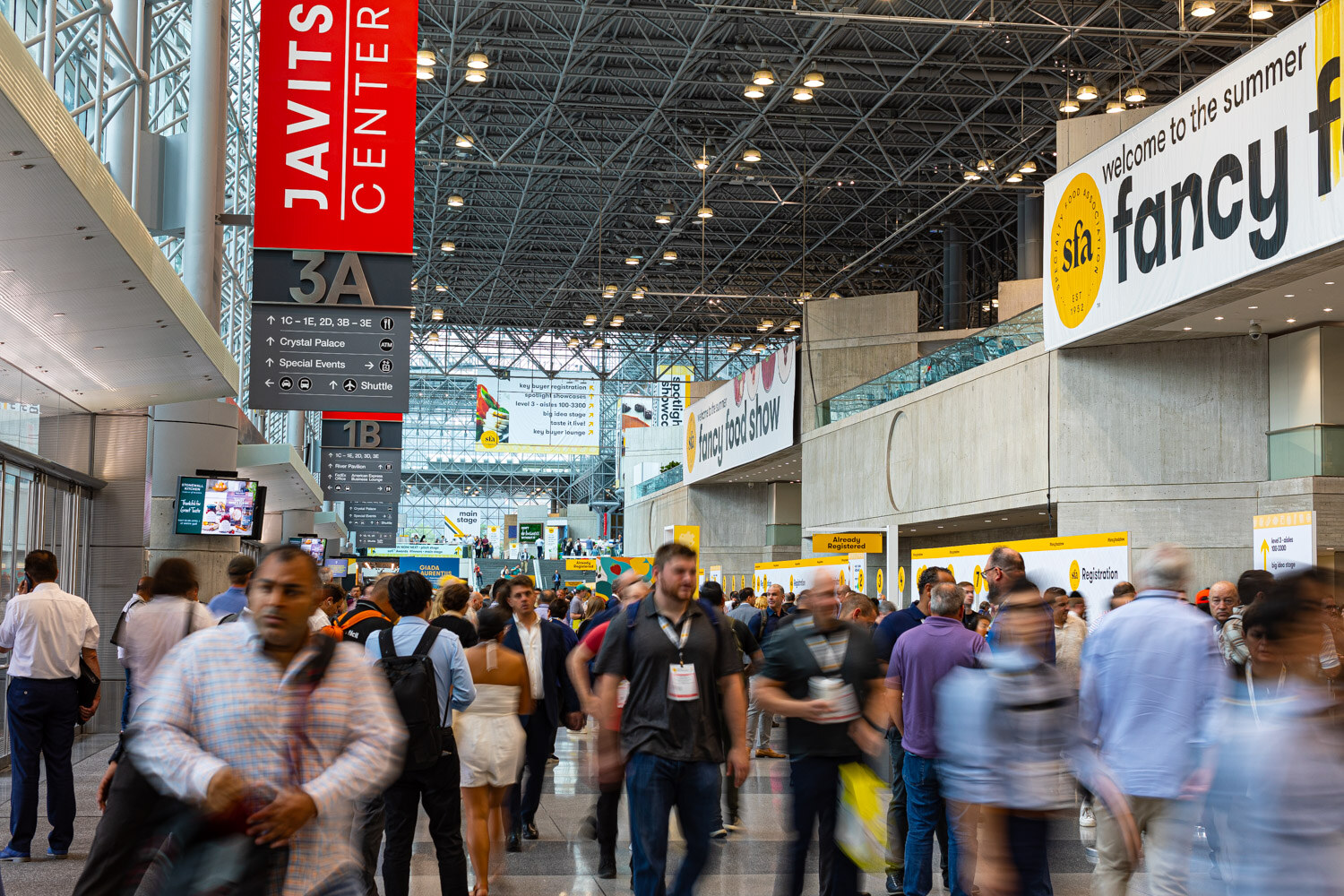 Motion-blurred attendees walk through the Javits Center concourse under a fancy food show banner and a Javits Center sign