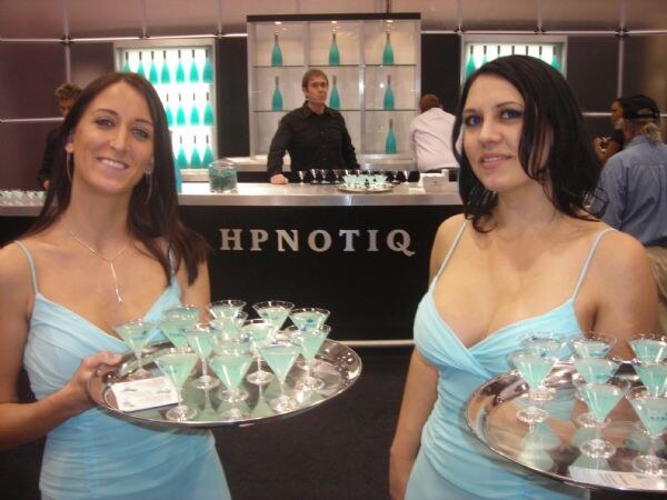 Two women in light blue dresses hold silver trays of pale blue-green drinks in martini glasses. Behind them, a man stands at a dark bar with the HPNOTIQ name and shelves of teal bottles.