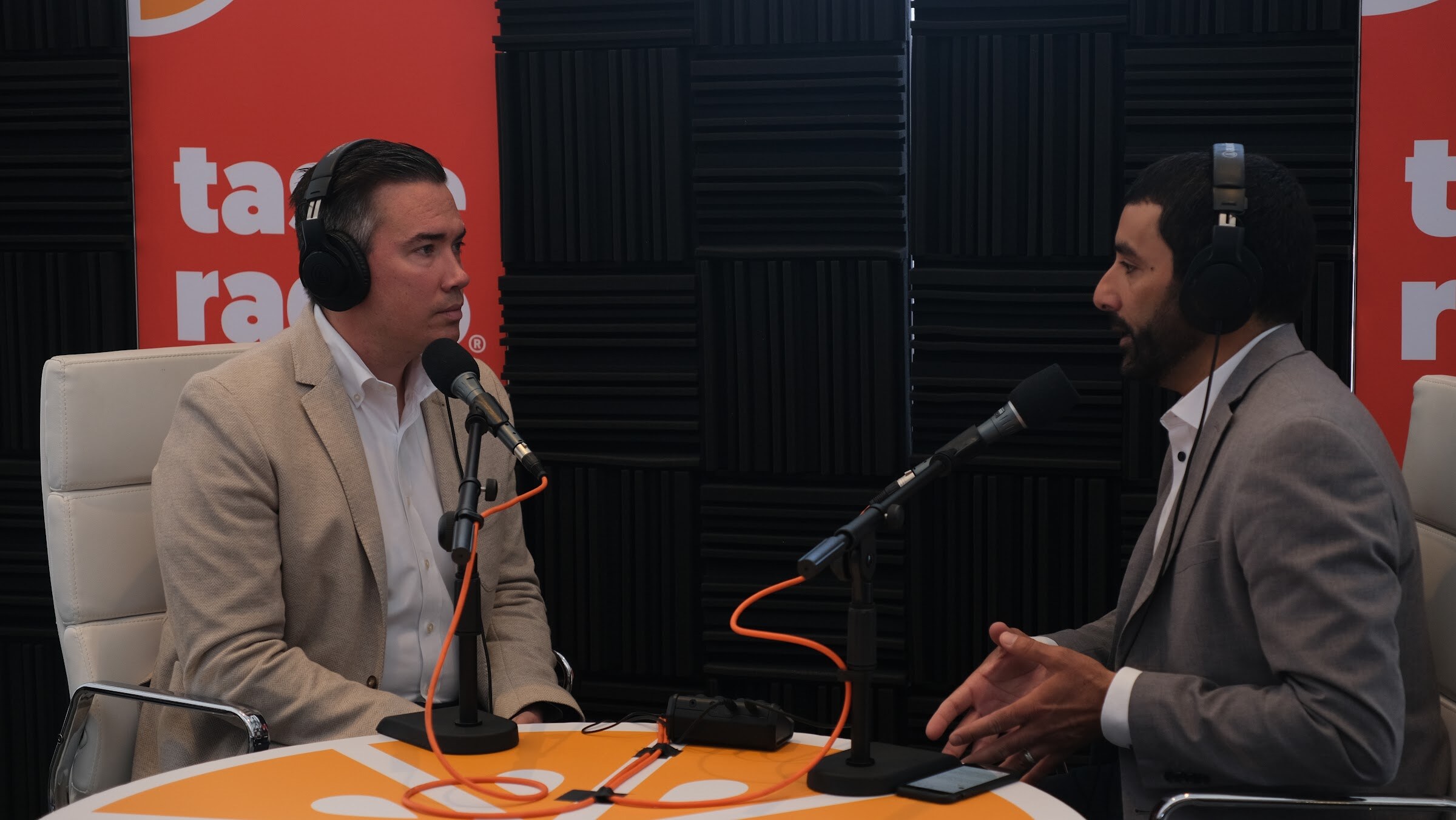 Man in a tan blazer wearing headphones at a microphone, facing a man in a gray blazer at an orange table in front of Taste Radio banners.