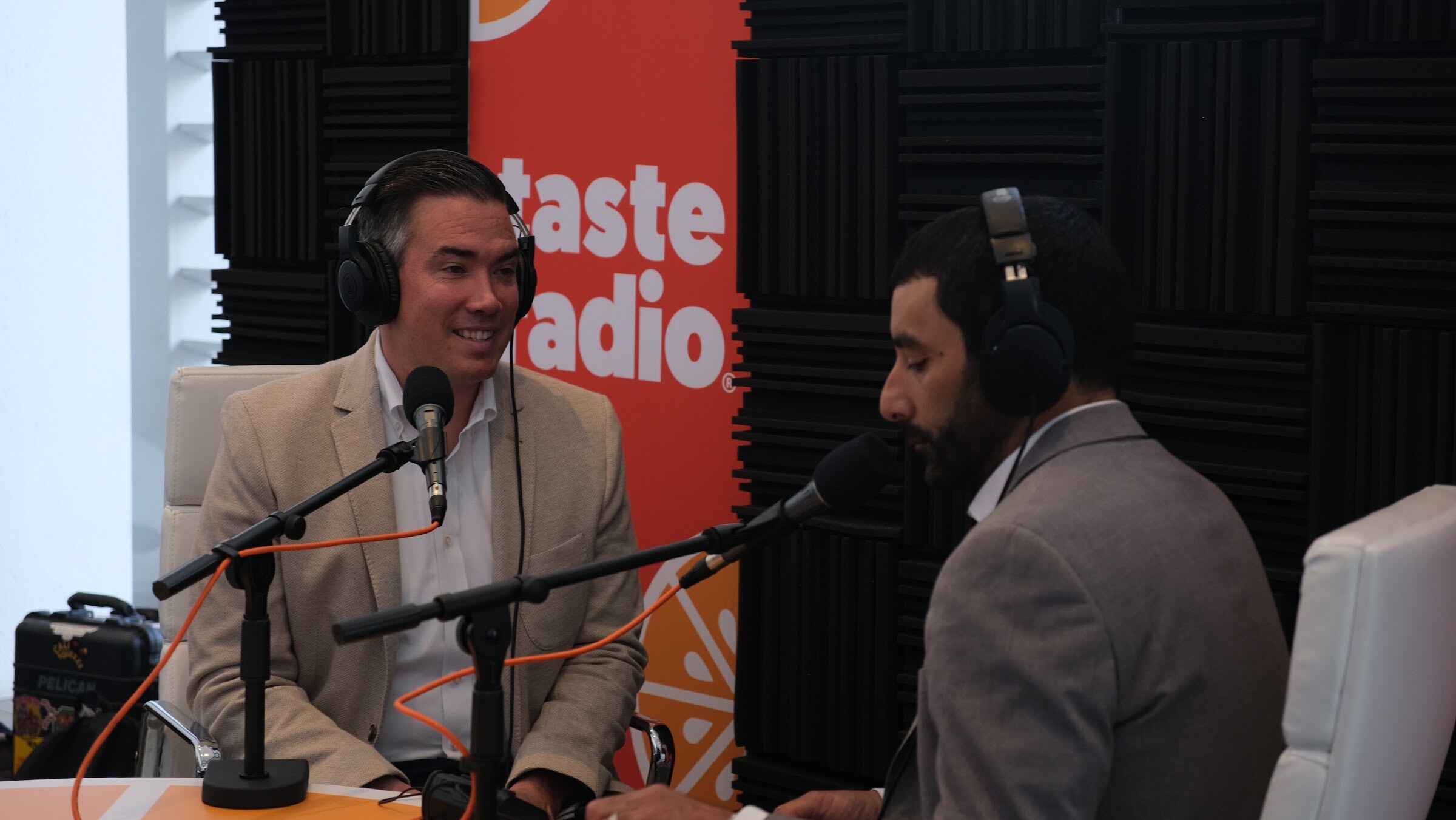 Smiling man in a tan blazer and headphones at a microphone, with the back of the interviewer at right and a Taste Radio banner behind.