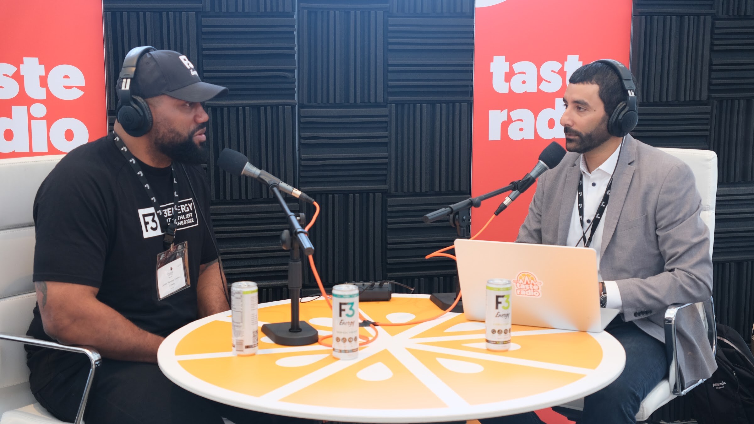 Man in a black F3 Energy shirt and cap wearing headphones across a round orange table with cans of F3 Energy, facing the interviewer and a laptop.
