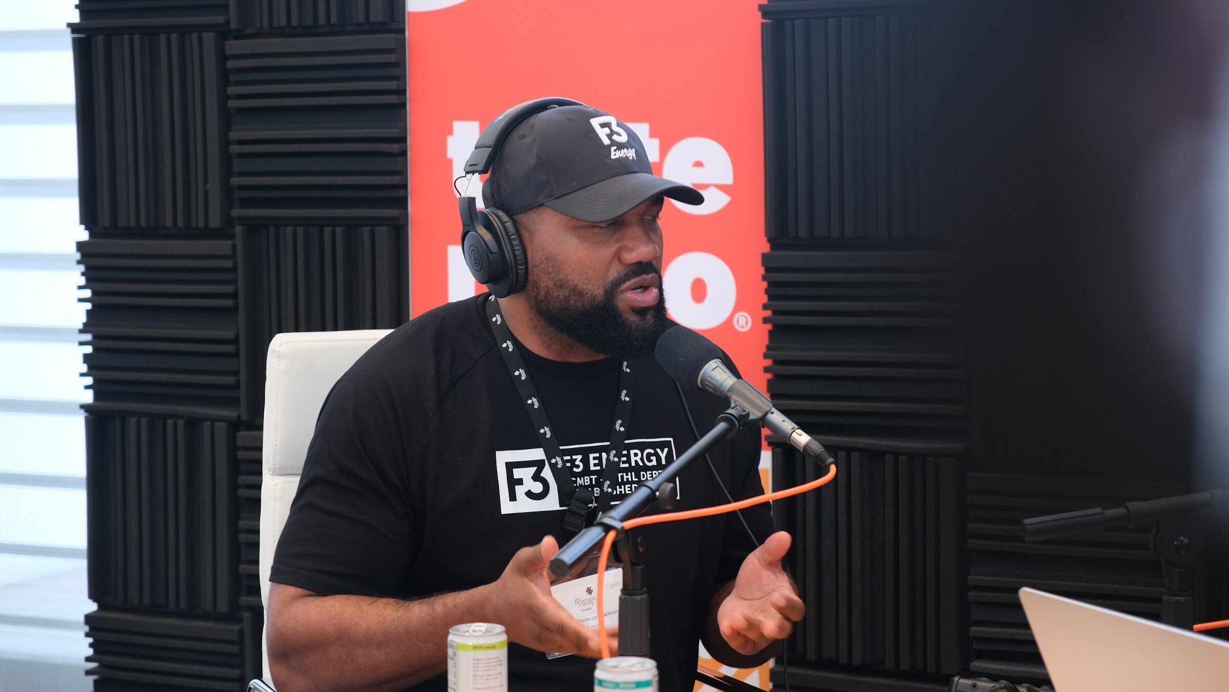 Man in a black F3 Energy shirt and cap with headphones, speaking into a microphone in front of black acoustic foam and a Taste Radio banner.