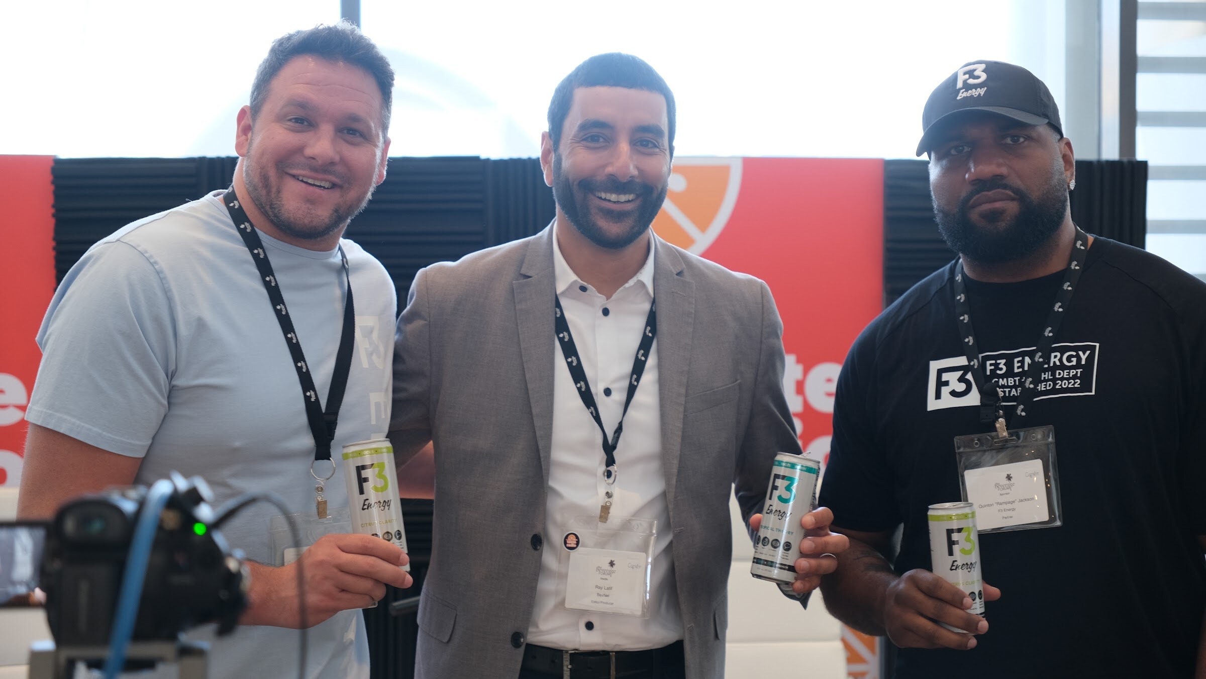Three men standing close together, two holding cans of F3 Energy, with Taste Radio banners behind them.