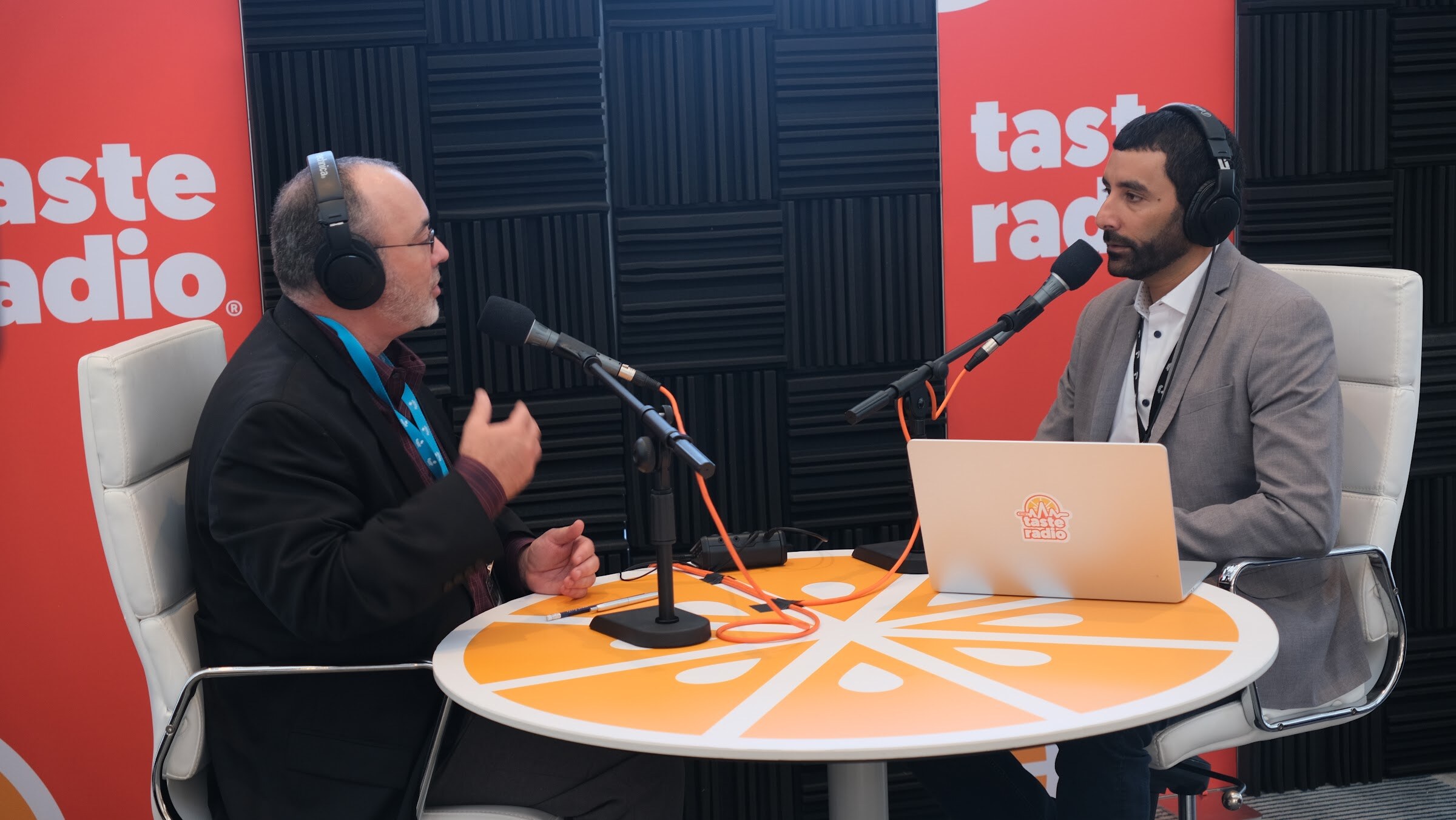 Man with glasses in a black jacket and headphones gesturing at a microphone across from the interviewer, with a laptop on the orange table.