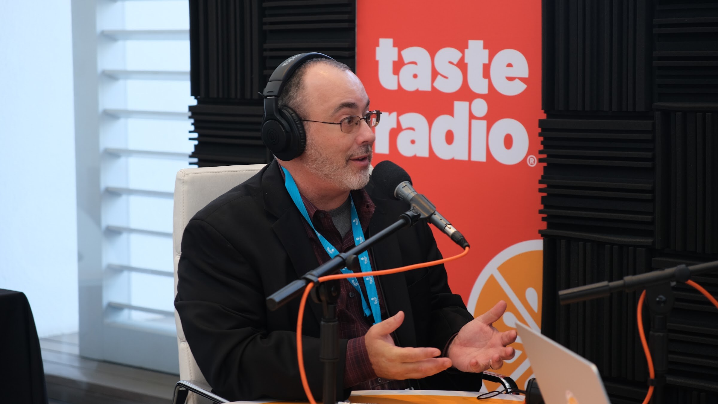 Bearded man with glasses, headphones and a conference lanyard speaking at a microphone with a Taste Radio banner behind him.