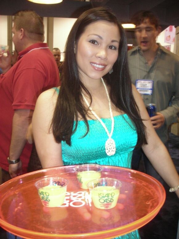 A smiling woman in a teal top and long white necklace holds an orange tray of small cups of yellow drink marked Island Oasis. Other attendees stand behind her.