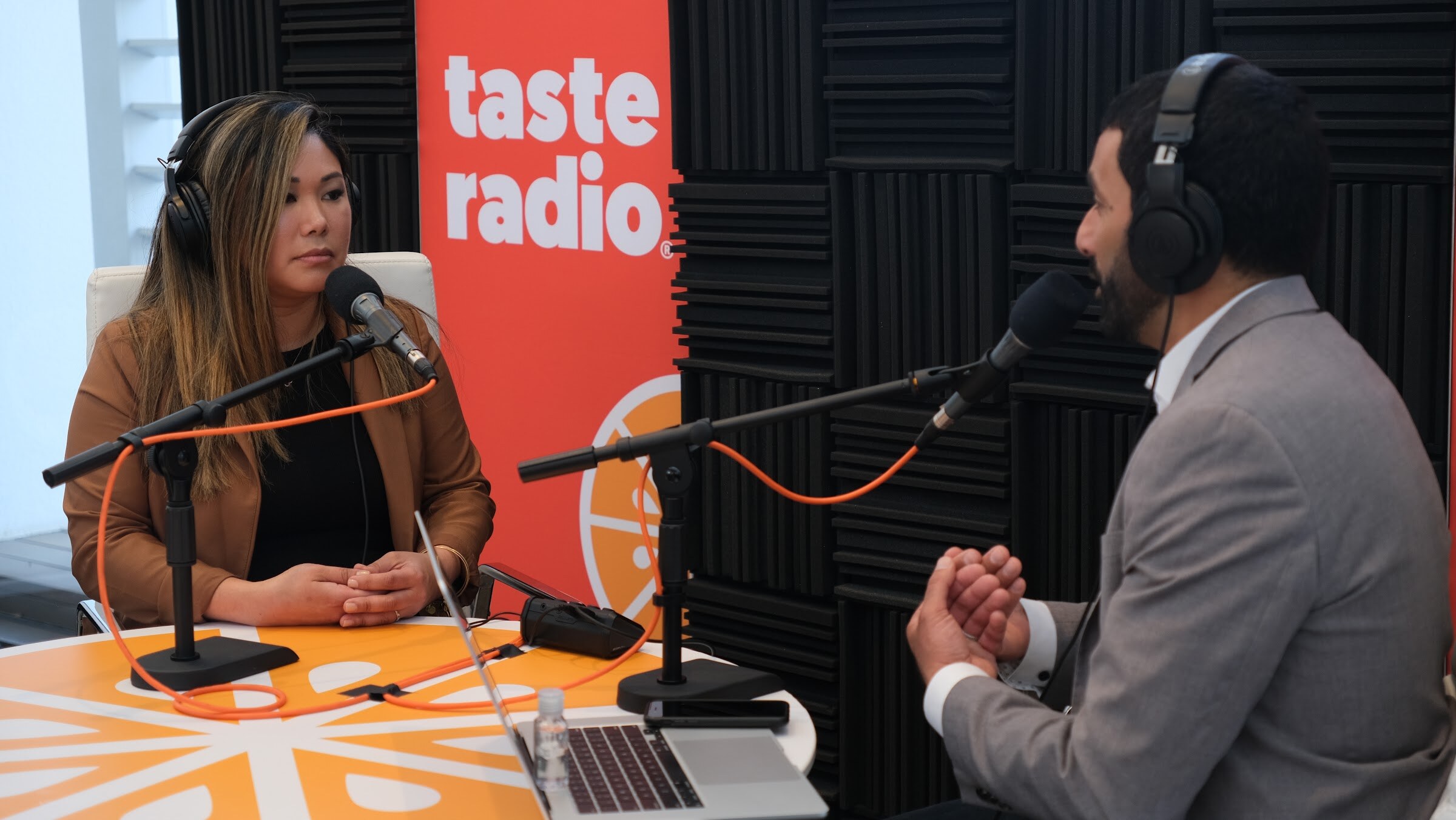 Woman in a brown jacket and headphones with hands folded at a microphone, facing the interviewer in a gray blazer.