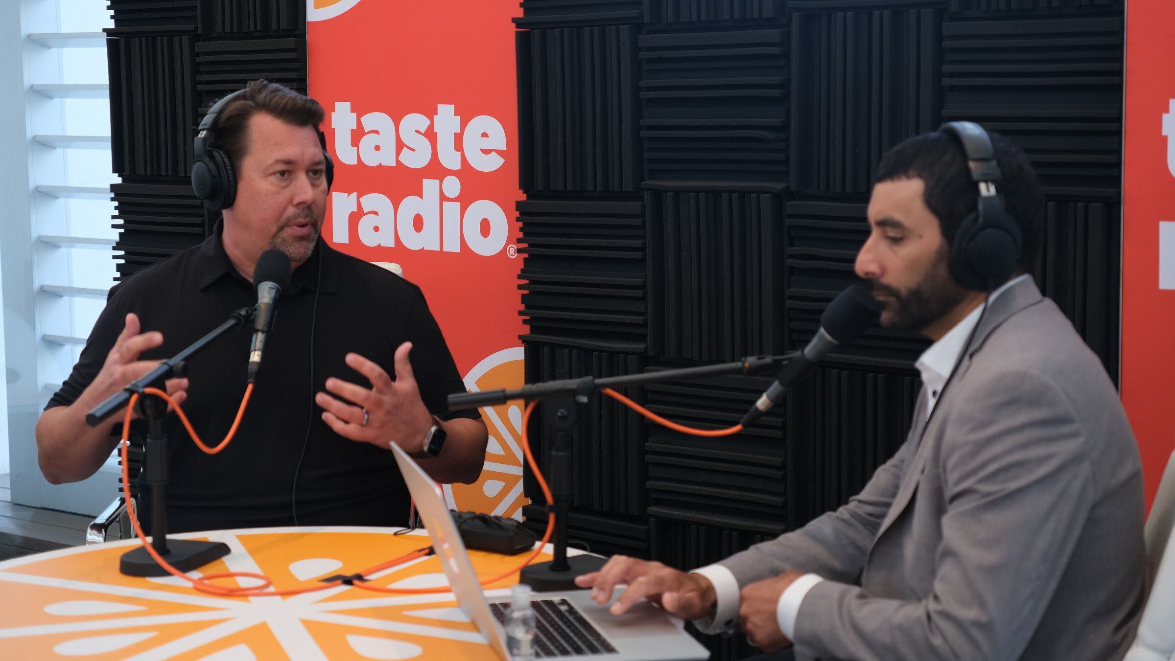 Man in a black polo and headphones gesturing at a microphone while the interviewer types on a laptop at the orange table.