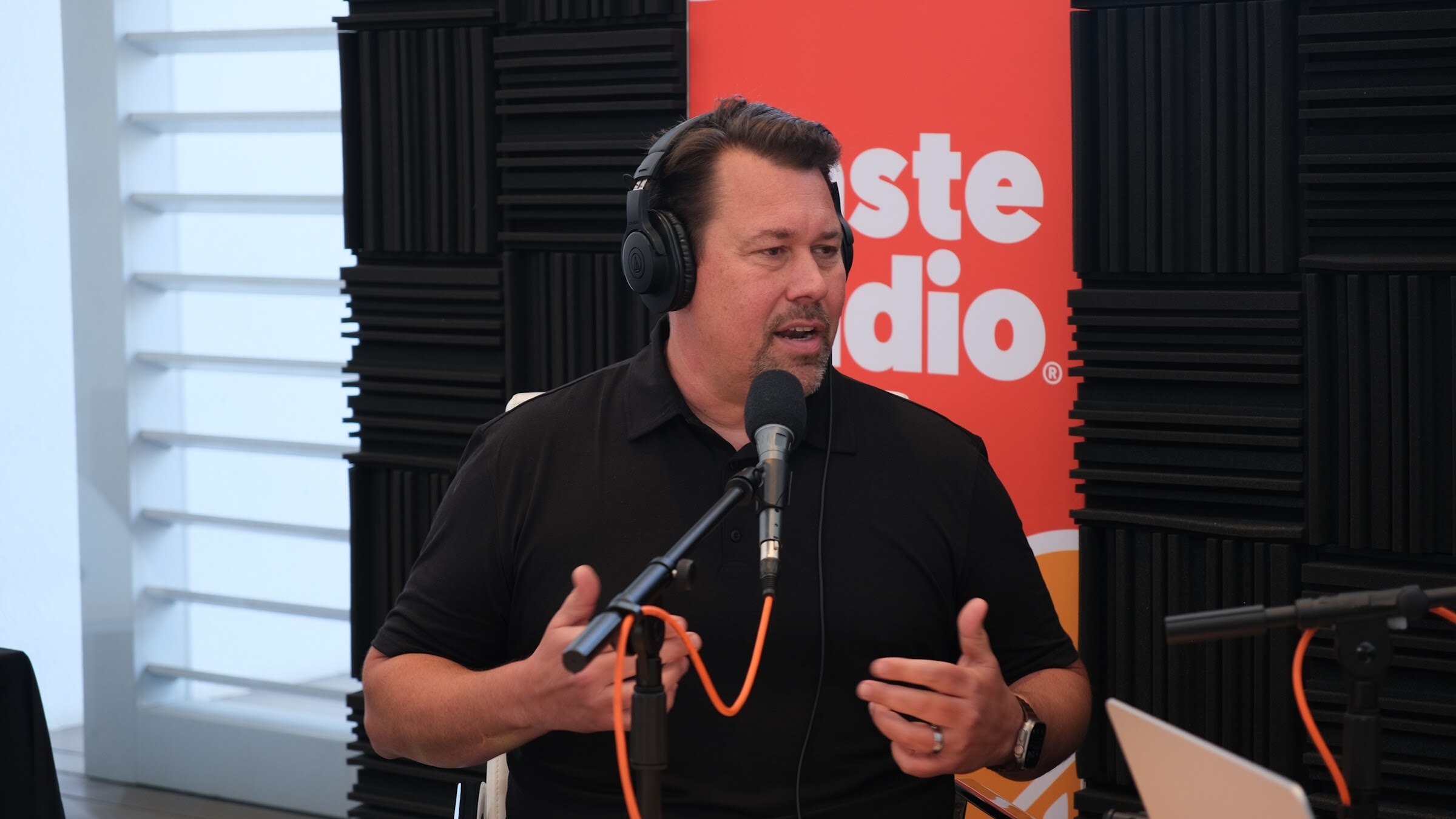 Man in a black polo and headphones speaking at a microphone in front of a Taste Radio banner and black foam panels.