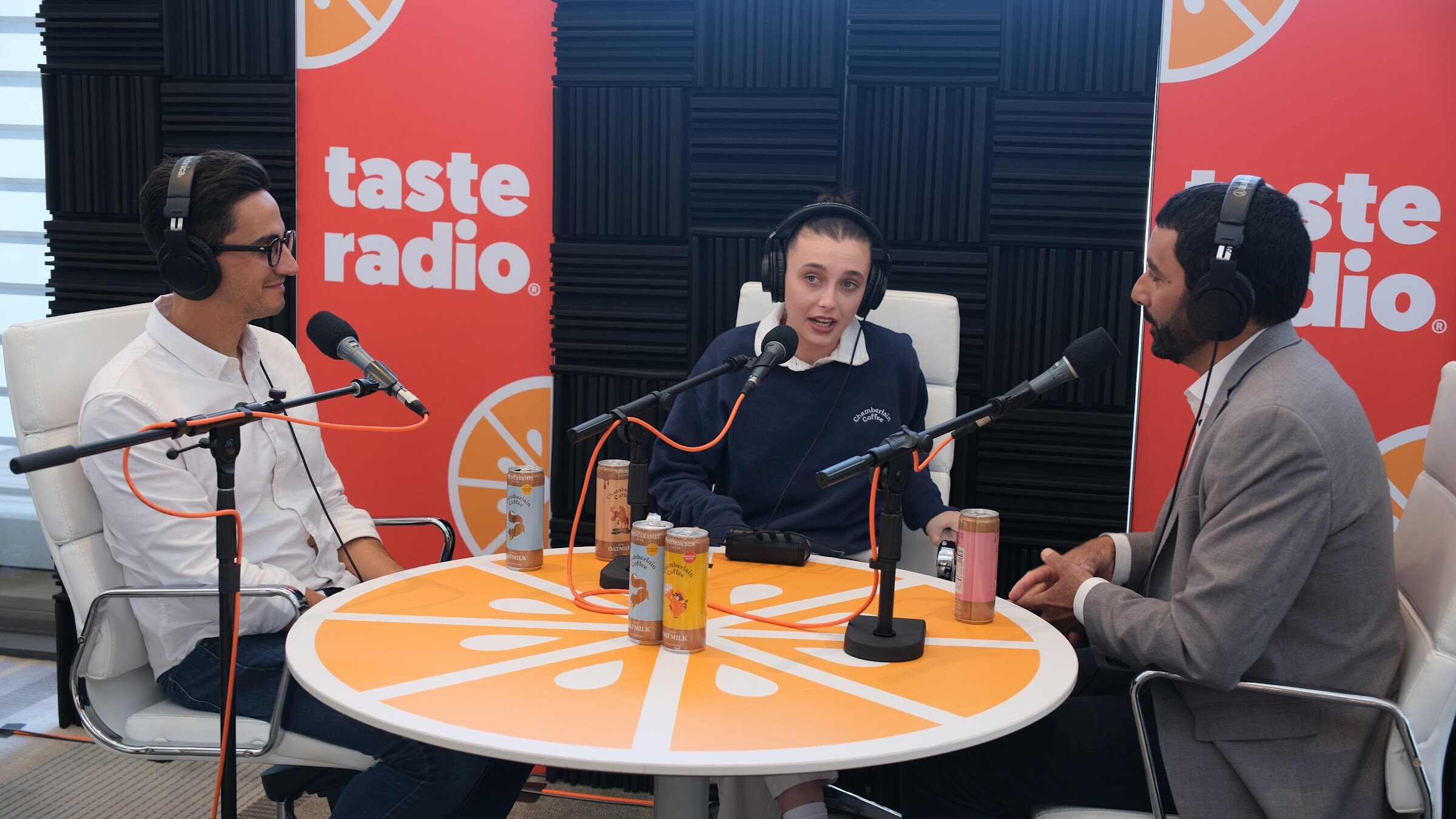 Two guests wearing headphones at a round orange table with several beverage cans, facing the interviewer.