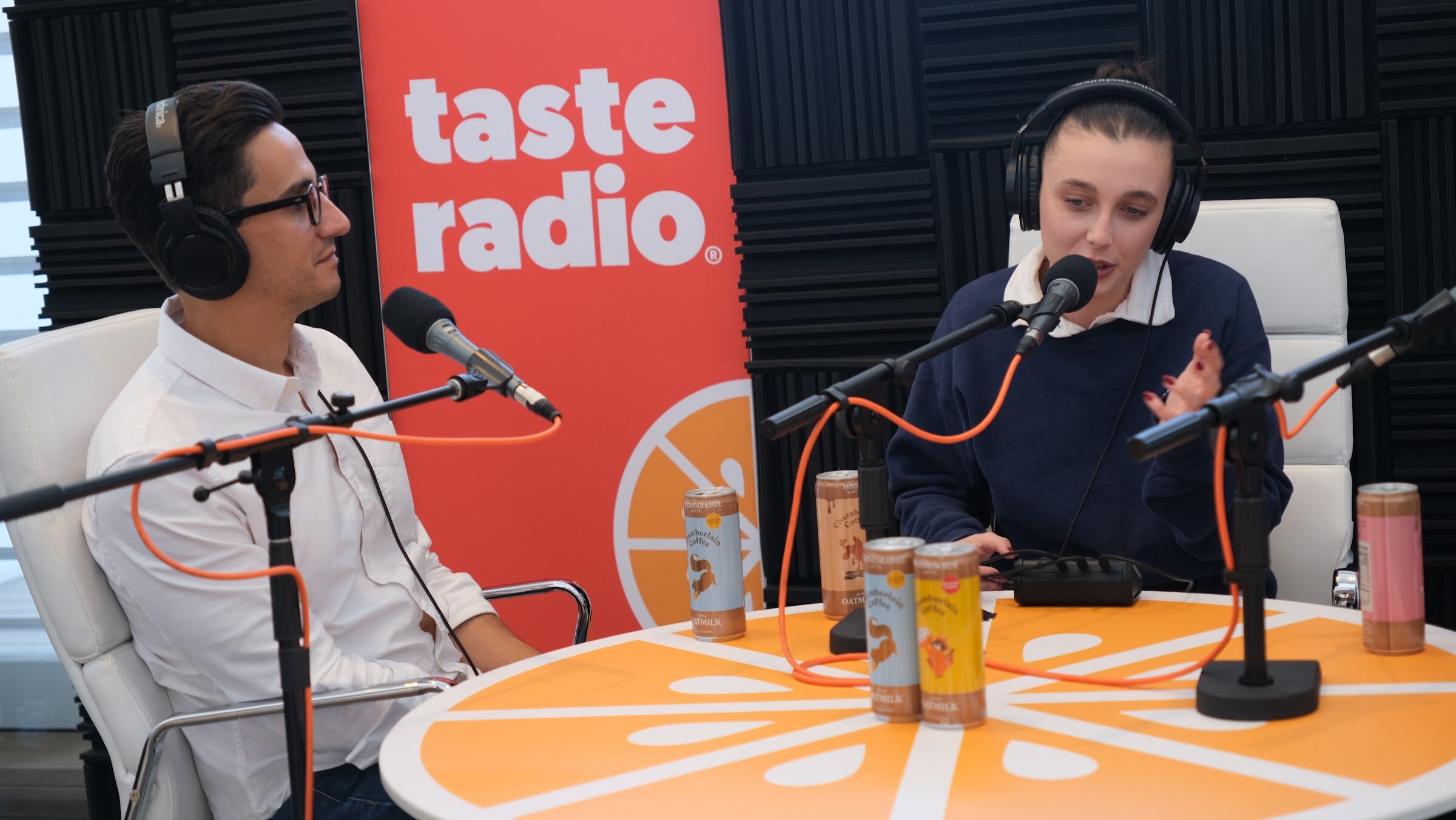 Man in a white shirt and a young woman in a navy sweatshirt, both in headphones, with cans on the table in front of a Taste Radio banner.