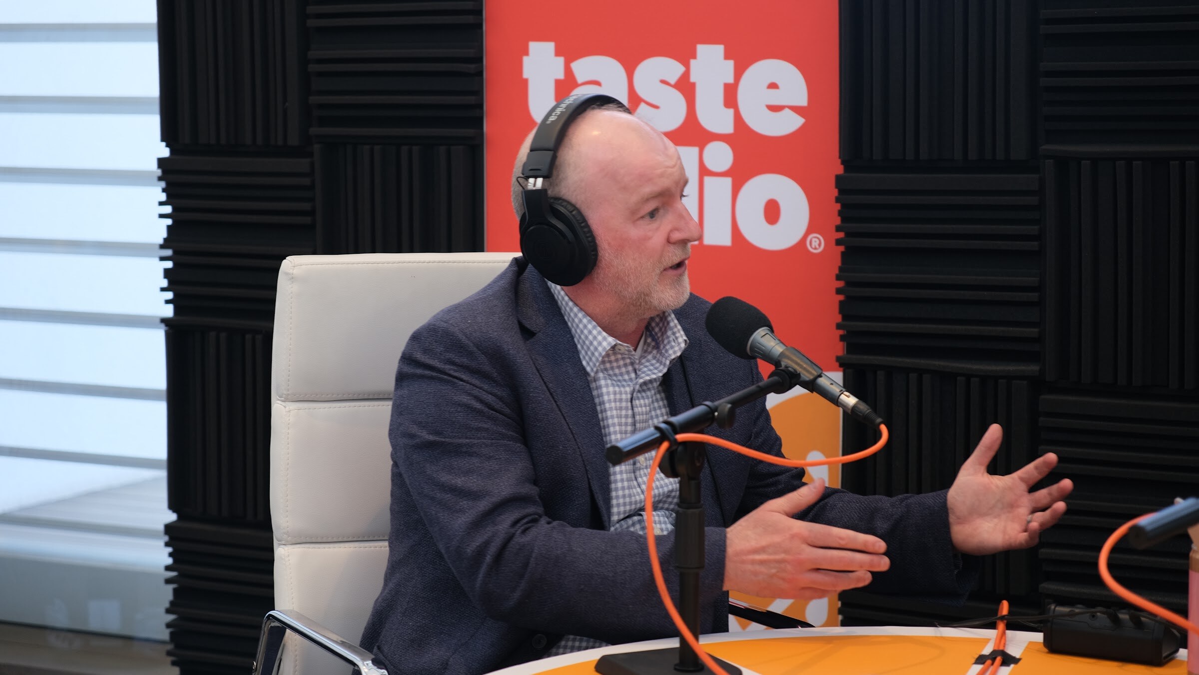 Gray-haired man in a navy blazer and headphones gesturing toward a microphone with a Taste Radio banner behind.