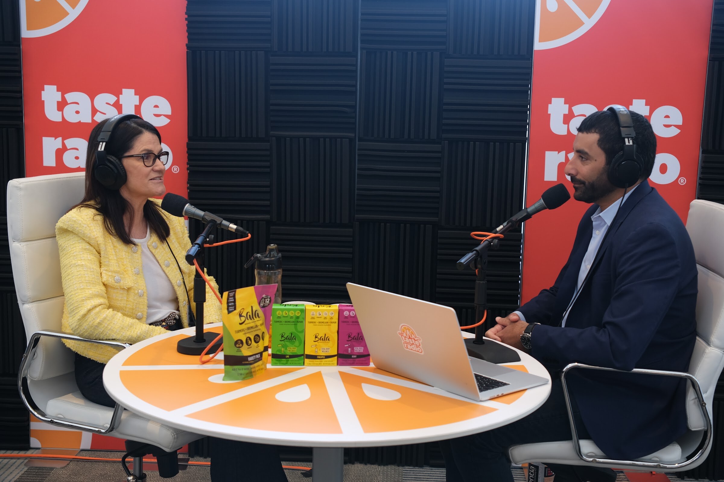 Woman in a yellow tweed jacket and headphones at a microphone, with Bala-branded packages on the orange table and the interviewer opposite.