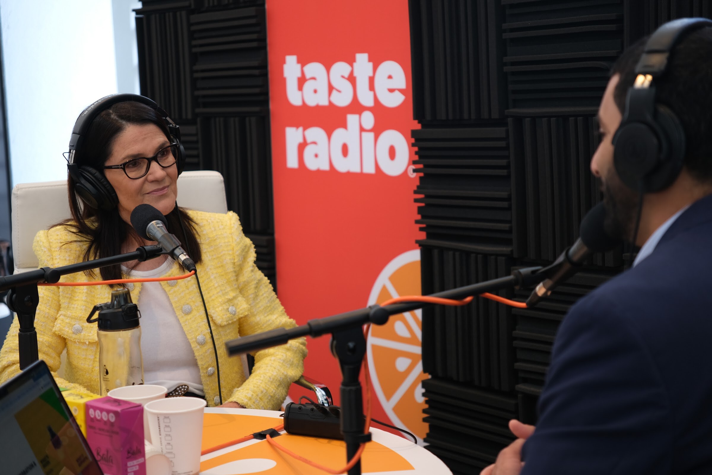 Woman with glasses in a yellow tweed jacket and headphones at a microphone, with the interviewer in the foreground.