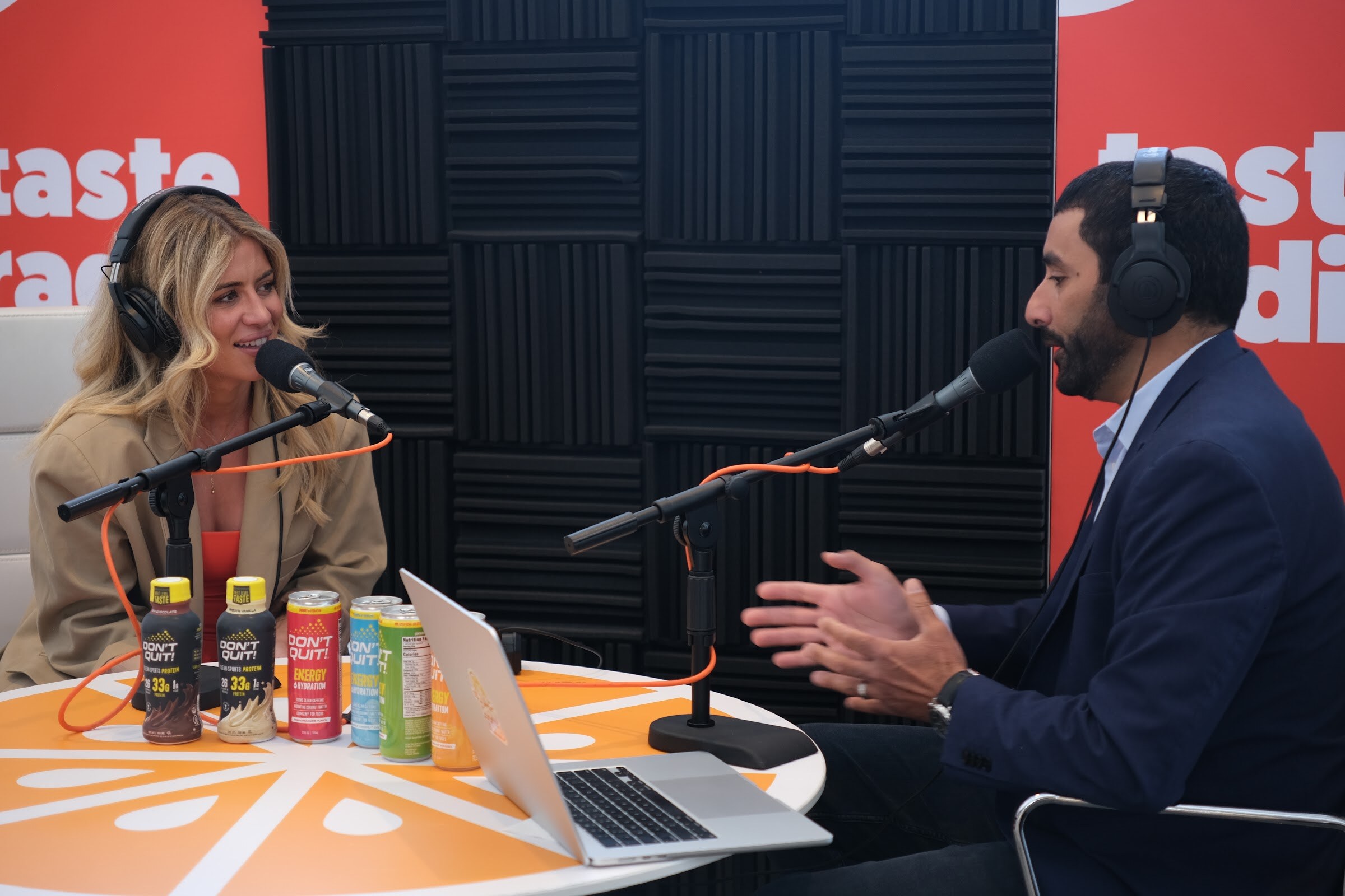 Blonde woman in a tan blazer headphones across the orange table from the interviewer, with small bottles and cans lined up on the table.