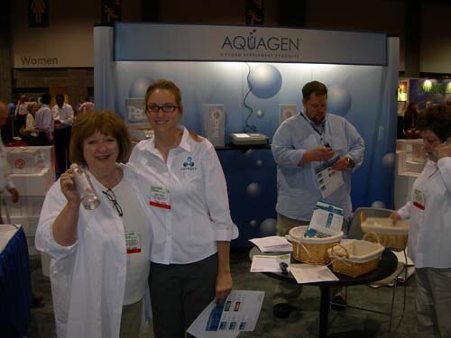 Two women in white coats stand at the Aquagen booth, one holding up a bottle; a man in a light blue shirt stands behind them near a table with brochures and baskets.