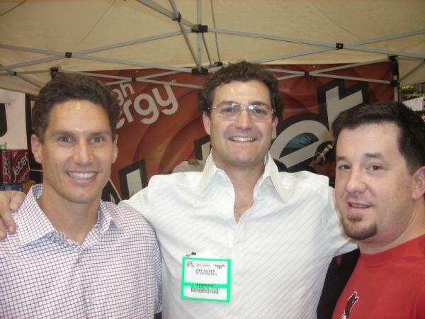 Jeff Silver, wearing a white striped shirt and an exhibitor badge, stands between two smiling men under a tent canopy with a dark red banner behind them.