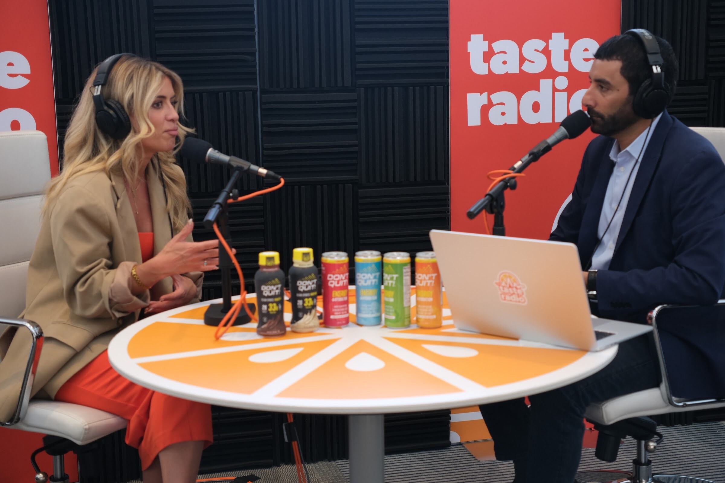 Woman in a tan blazer and headphones speaking at a microphone with a row of cans and bottles on the orange table beside a laptop.
