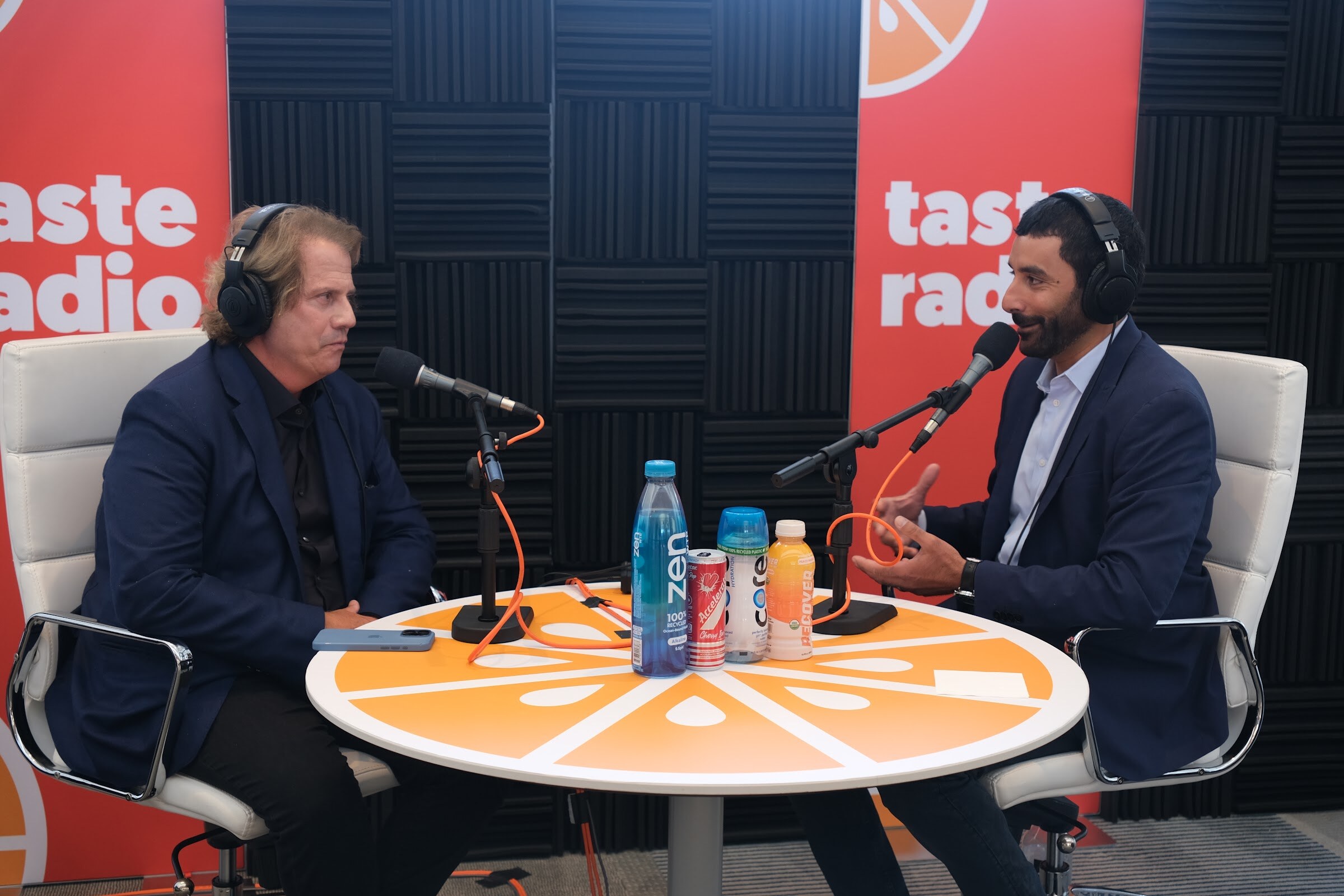 Man with long hair in a navy jacket and headphones across the round table from the interviewer, with bottles including Zen and Core on the table.