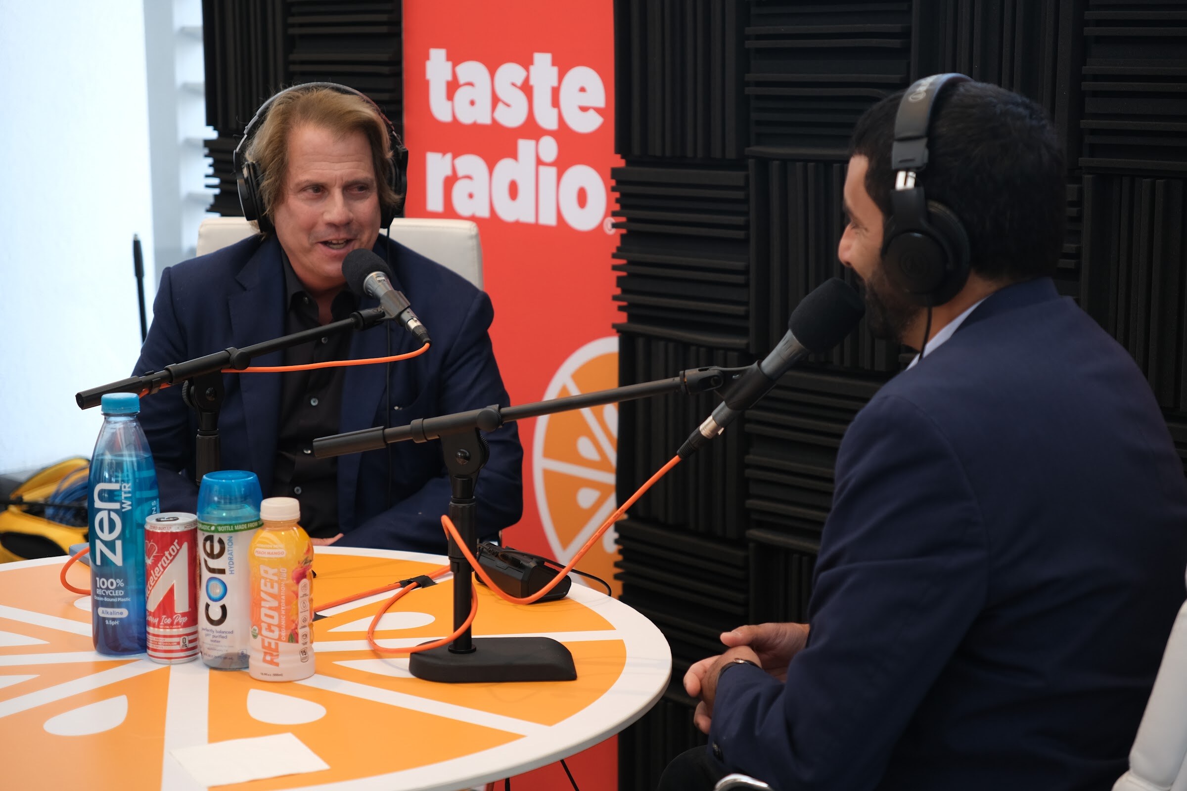 Smiling man in a navy blazer and headphones at a microphone with Zen and Core bottles on the table and the interviewer in the foreground.