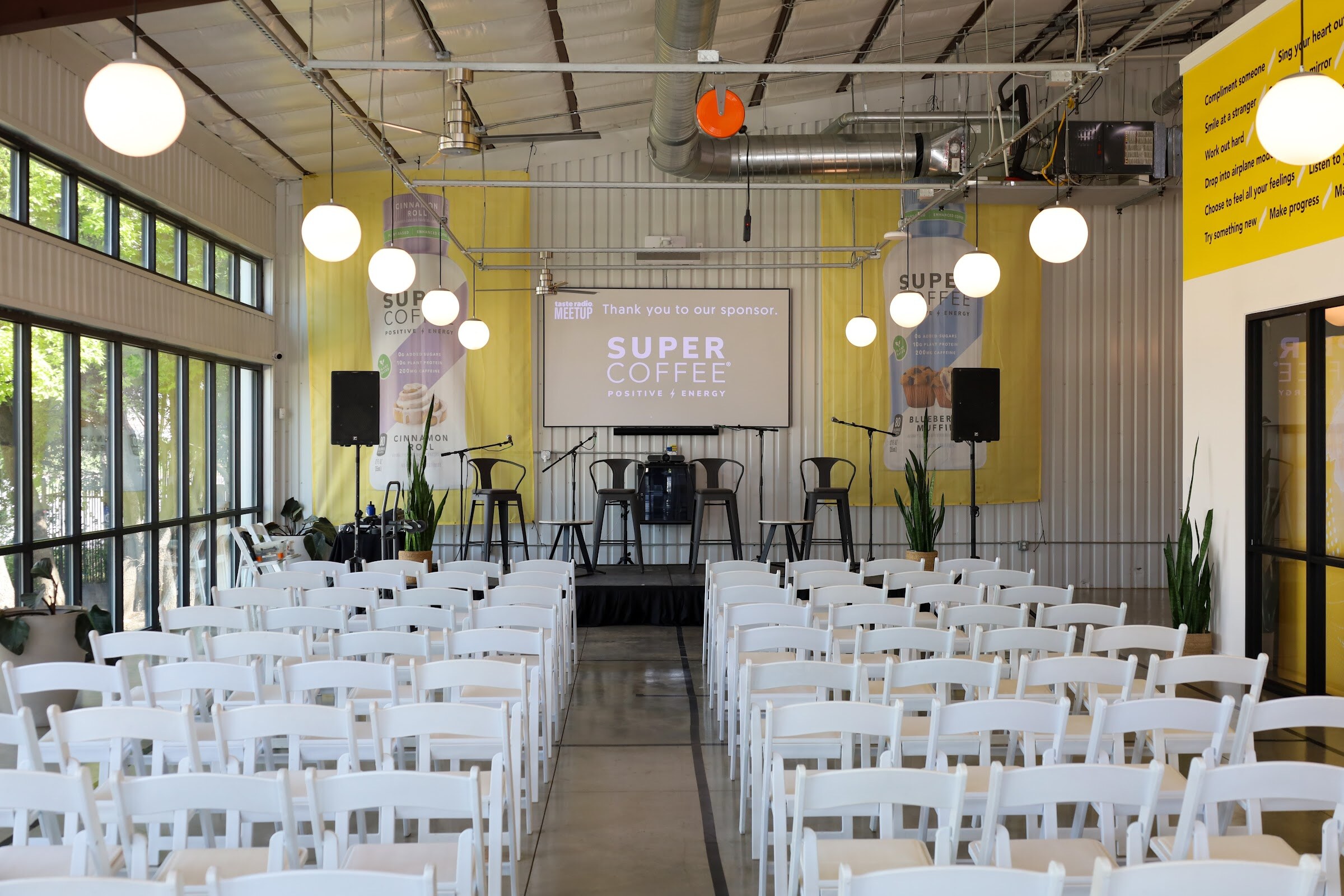 View down aisles of white folding chairs to a small stage with gray bar stools and microphone stands under a screen reading Taste Radio Meetup, Thank you to our sponsor, Super Coffee, between yellow Super Coffee banners, with round pendant lights overhead and tall windows at the left.