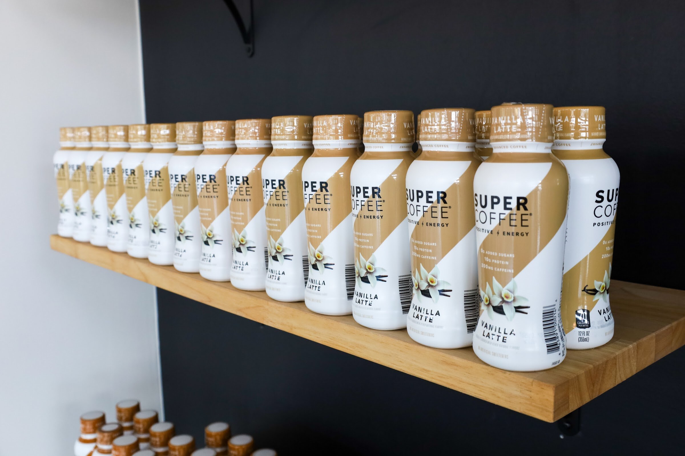 A row of white and tan Super Coffee Vanilla Latte bottles on a light wooden shelf against a black wall, with more bottles below.