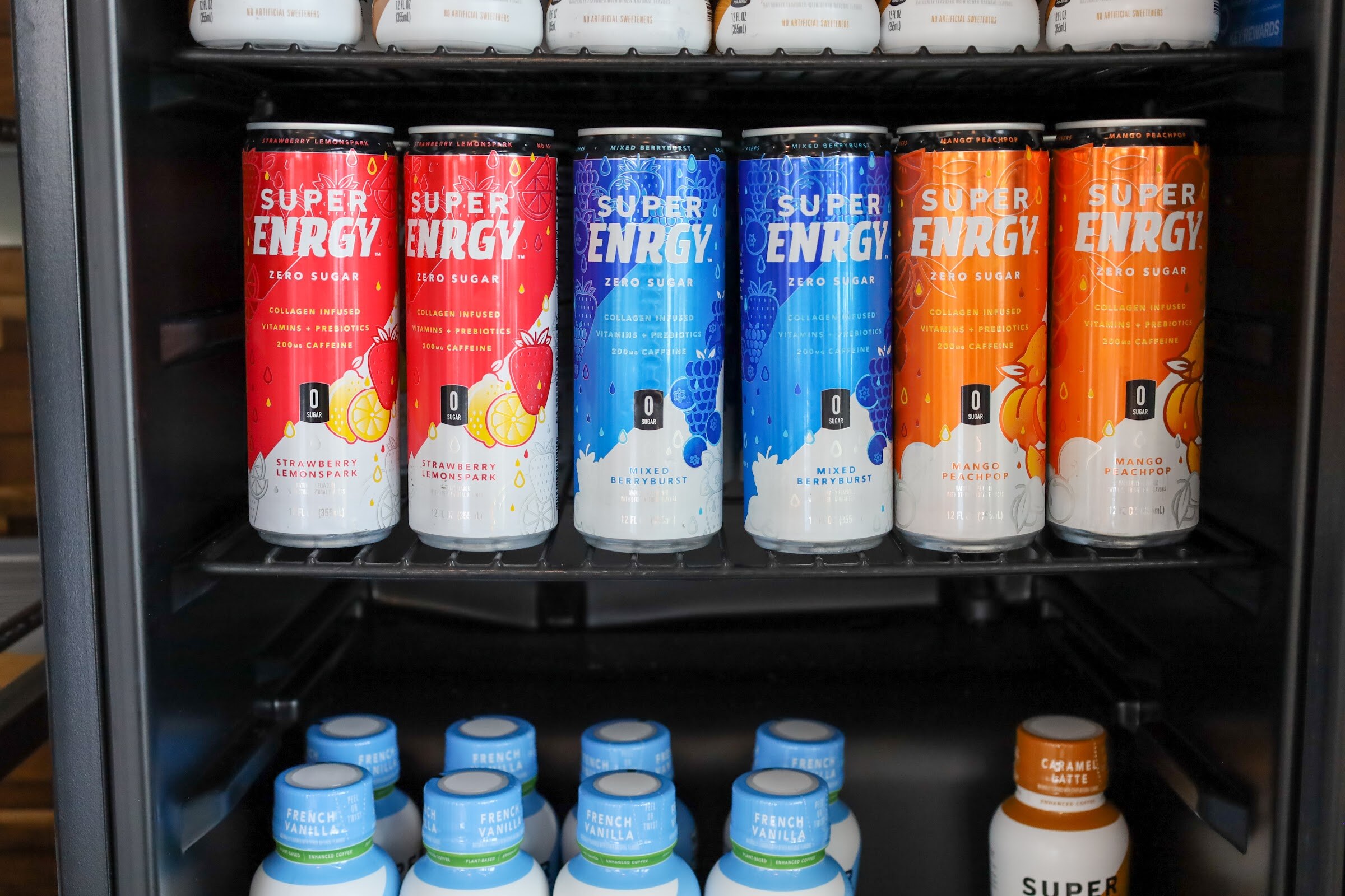 Pairs of red, blue and orange Super Enrgy zero sugar cans on a fridge shelf above bottles of French Vanilla and Caramel Latte Super Coffee.