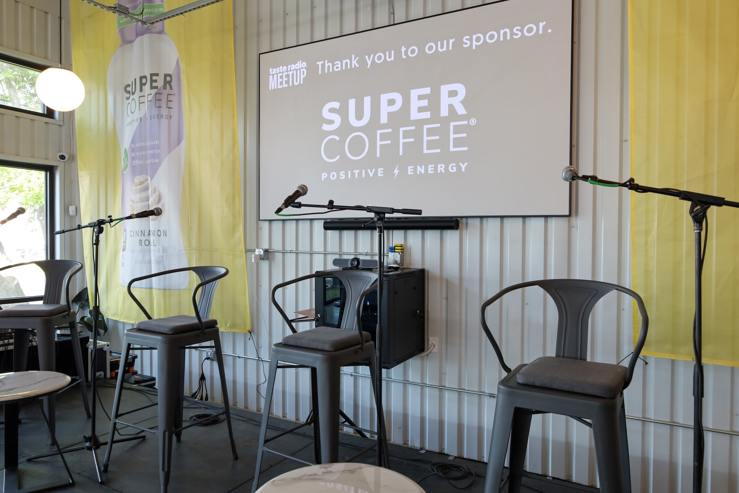 Four empty gray metal bar stools and microphone stands in front of a screen reading Taste Radio Meetup, Thank you to our sponsor, Super Coffee, between yellow Super Coffee banners.