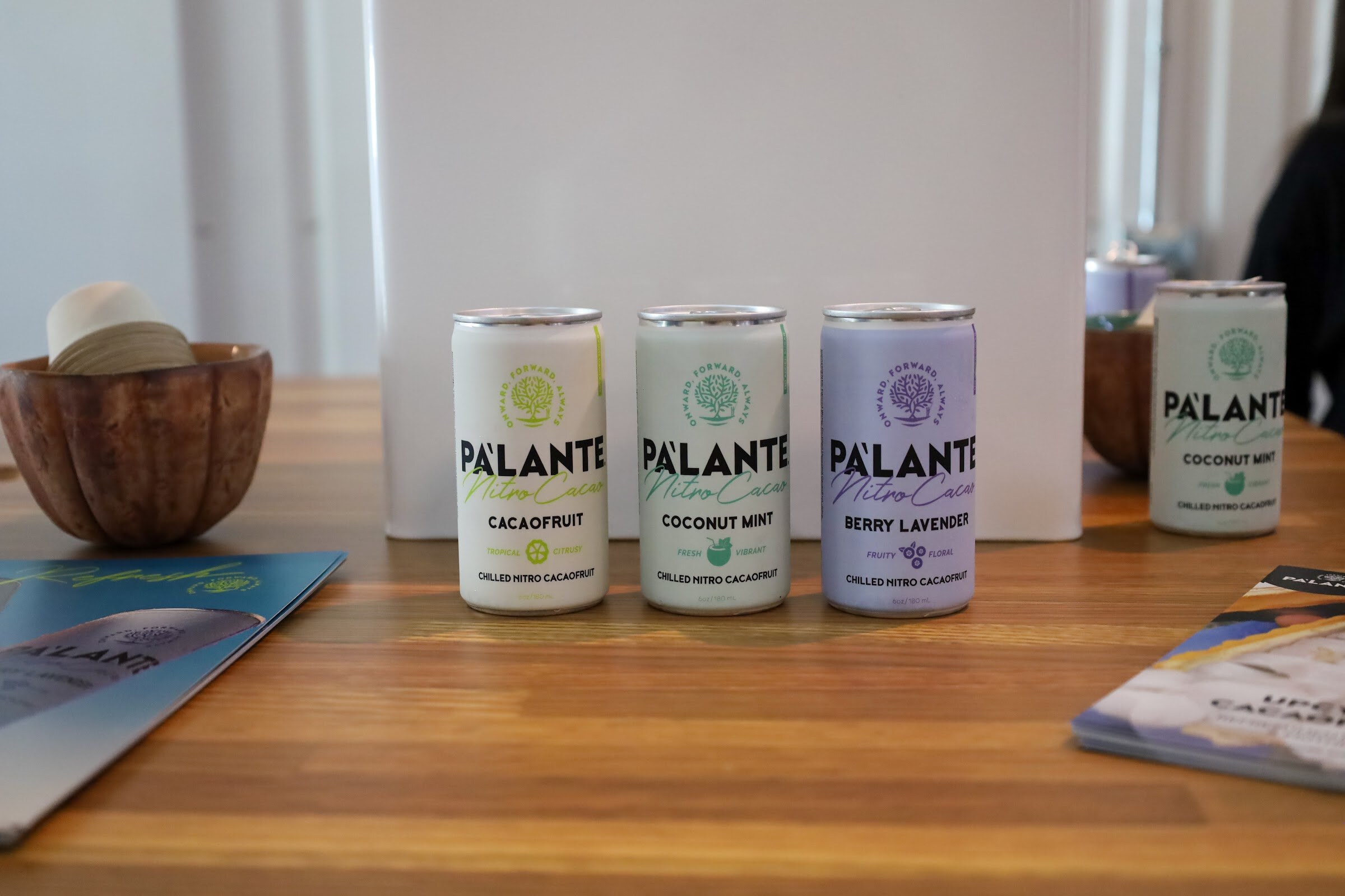 Cans of Pa'lante Nitro Cacao in Cacaofruit, Coconut Mint and Berry Lavender on a wooden table beside a wooden bowl and brochures.