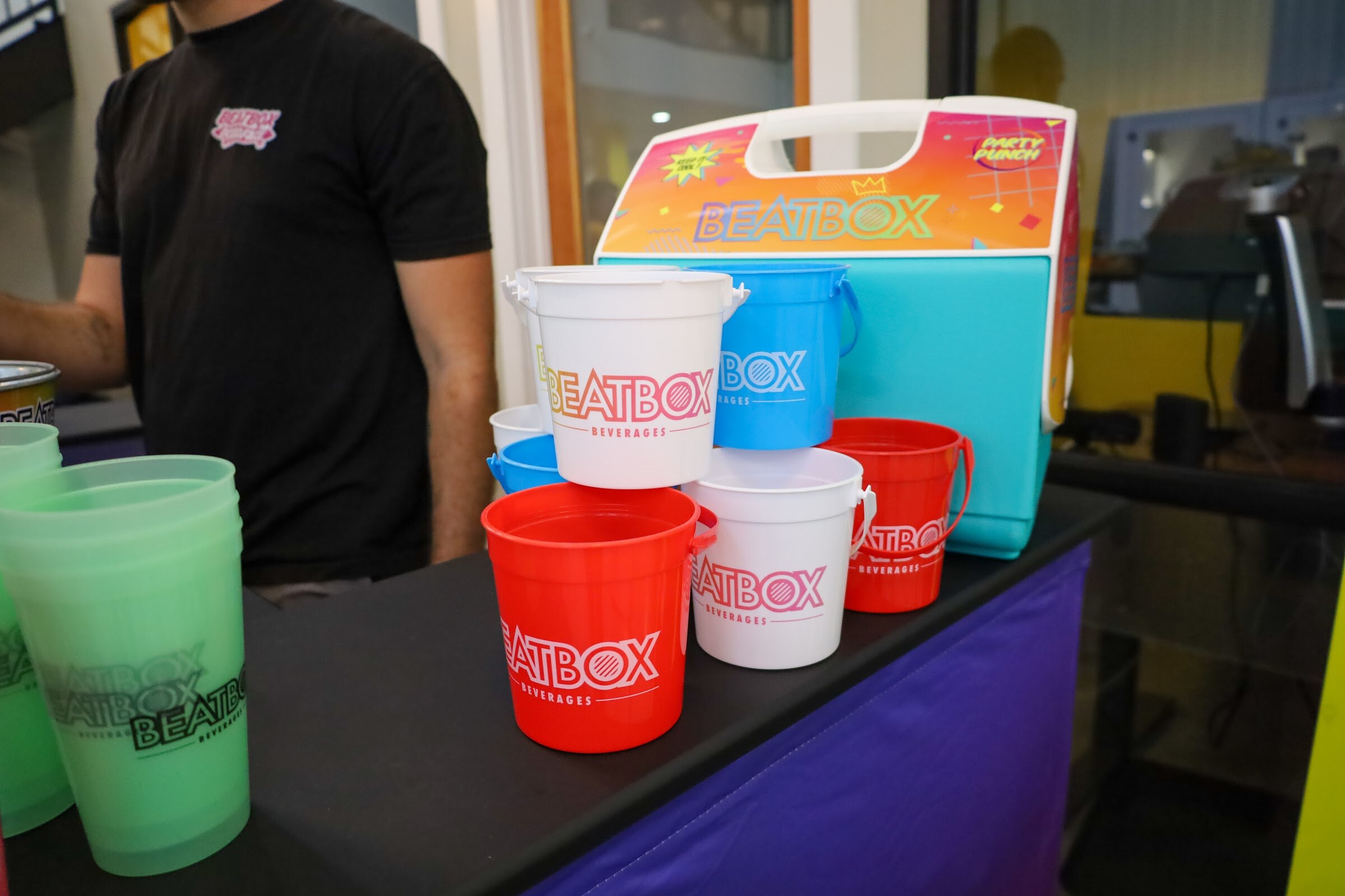 Stacked red, white and blue Beatbox Beverages buckets and green cups beside a turquoise Beatbox Party Punch cooler on a black table with a purple skirt, with a man in a black t-shirt standing behind it.