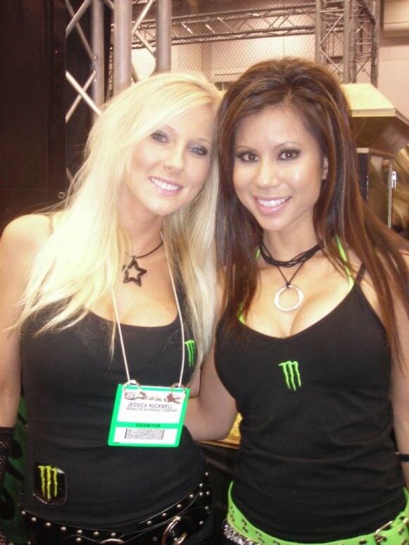 Two smiling women in black Monster Energy tank tops pose close together. The woman on the left wears an exhibitor badge reading Jessica Rockwell, Monster Beverage Company.