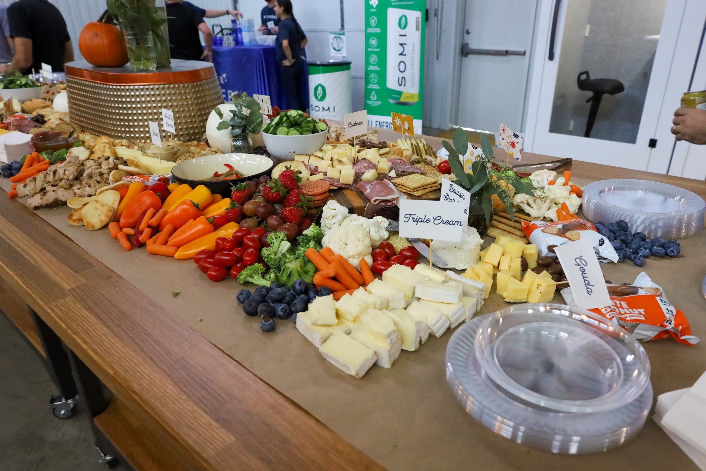 A long wooden table covered with brown paper holds cheeses labeled Triple Cream and Gouda, carrots, peppers, broccoli, cauliflower, berries and crackers, with a Somi banner and a blue-draped table at the back.