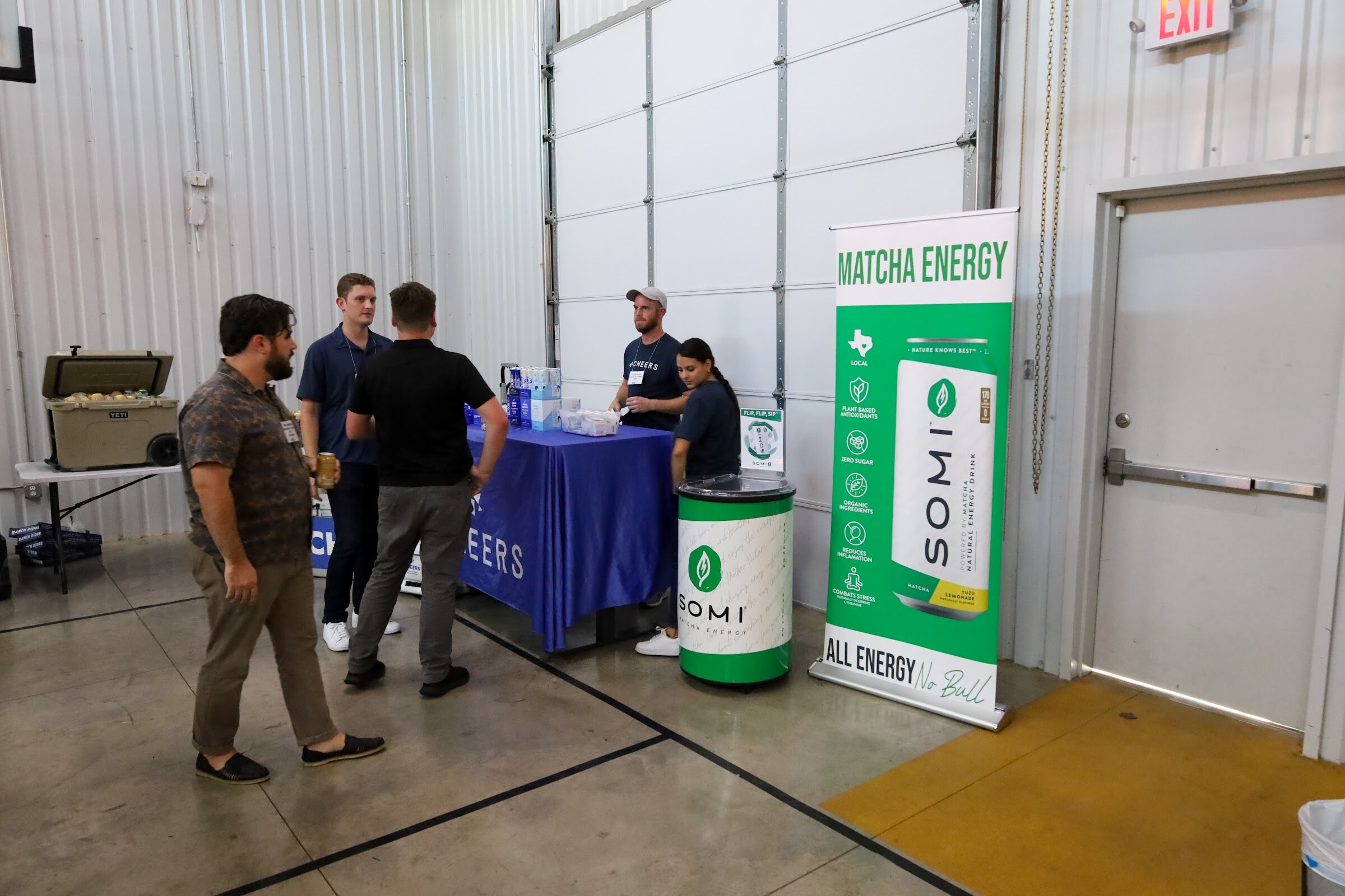 A tall banner reading Matcha Energy, Somi, All Energy No Bull stands beside a Somi cooler and a blue-draped table where two people in navy shirts stand, with three guests in front and a cooler at the left.