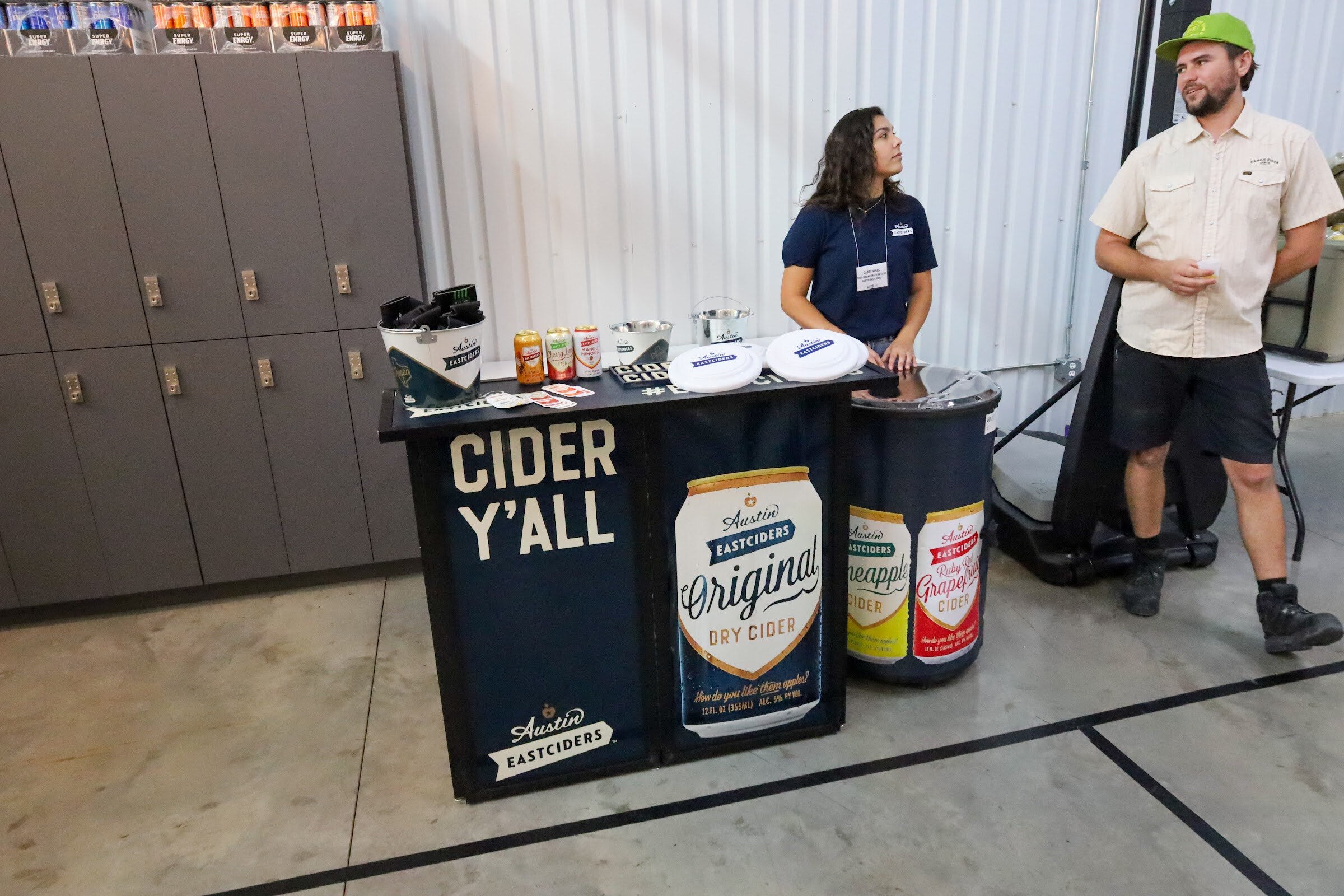 A black table front reading CIDER Y'ALL with an Austin Eastciders Original Dry Cider can graphic, a woman in a navy shirt behind it and a man in a green cap and tan button-up shirt beside it, with gray lockers and boxes on a shelf behind them.