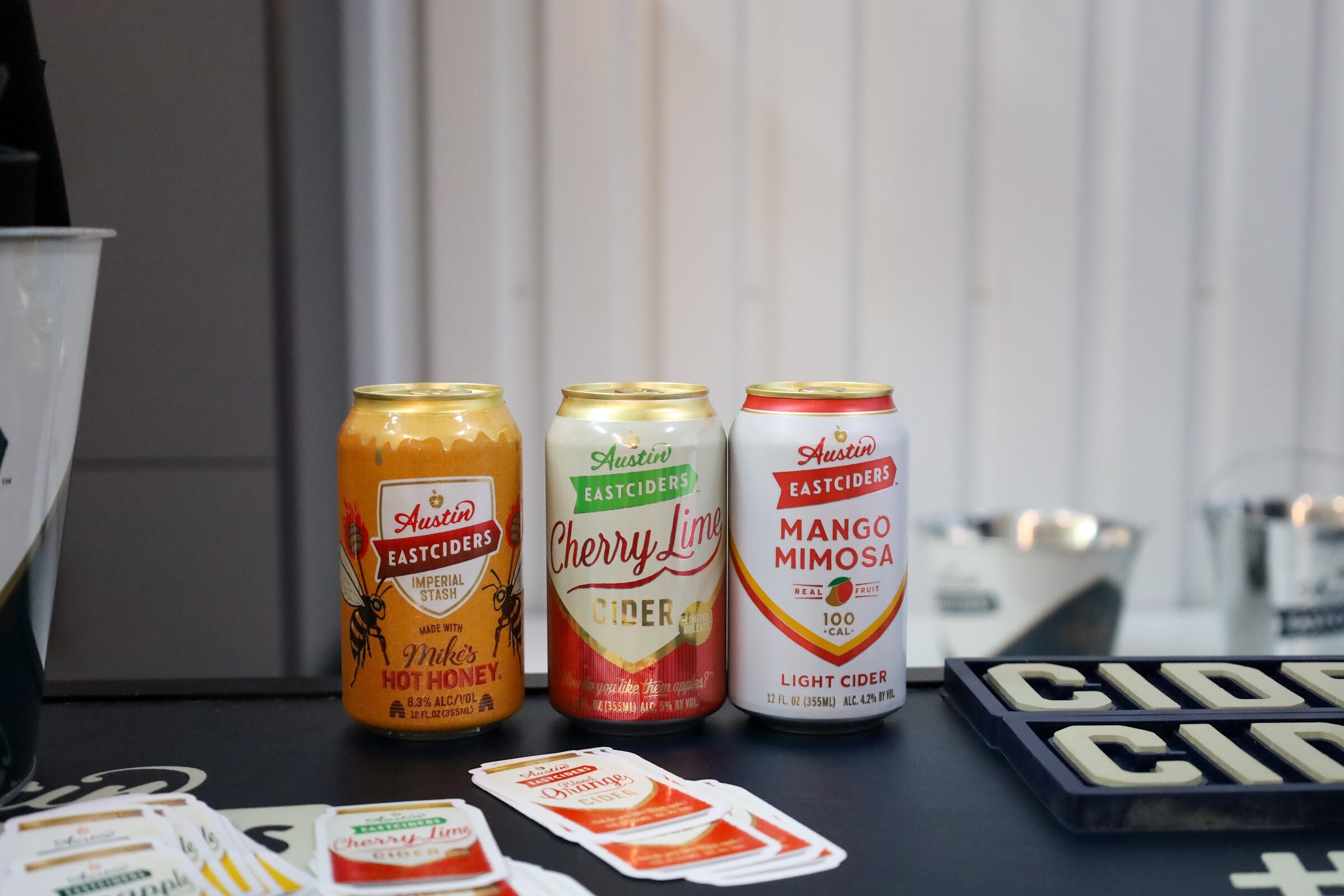 Three Austin Eastciders cans, Imperial Stash Made with Mike's Hot Honey, Cherry Lime Cider and Mango Mimosa Light Cider, stand on a black table with sample cards.