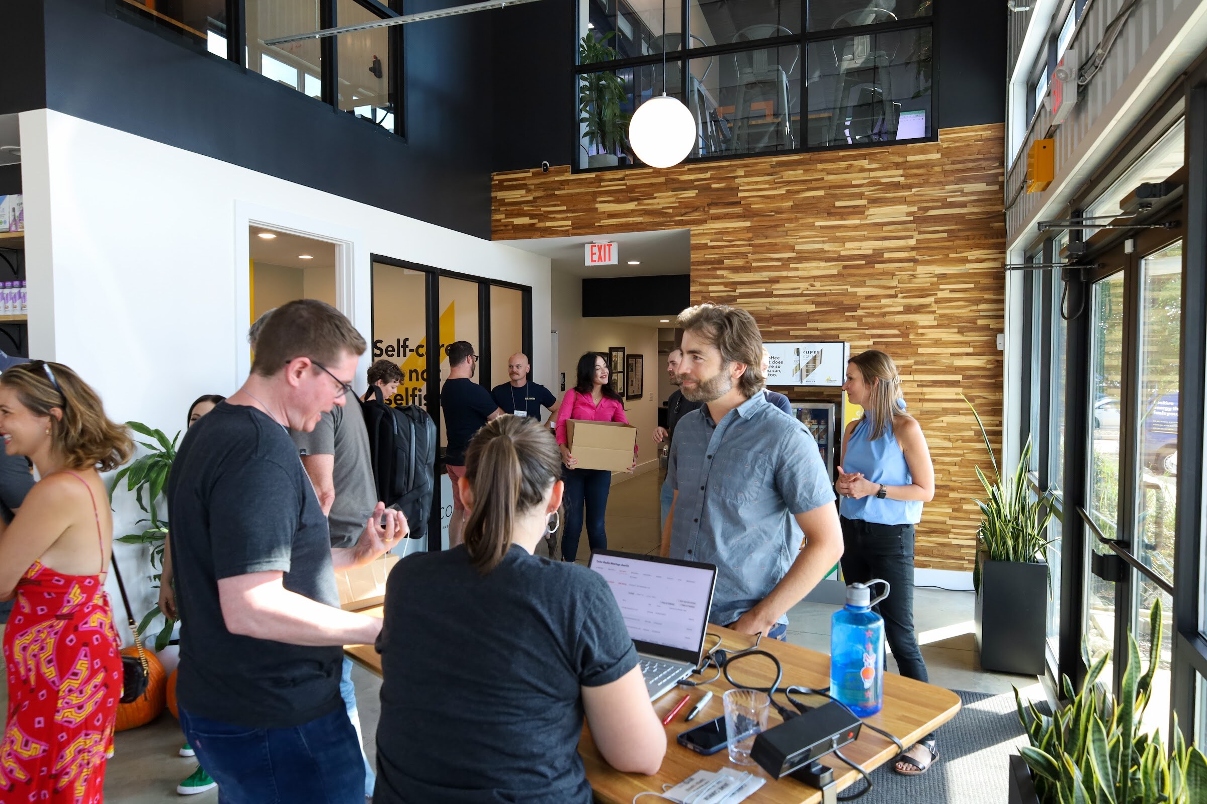 Guests gather around a light wooden table with a laptop, a label printer and a blue water bottle in a bright two-story entry lobby with a wood-paneled wall, a woman in a black t-shirt at the laptop, a man in a gray shirt facing her and a bearded man in a gray-blue shirt waiting.