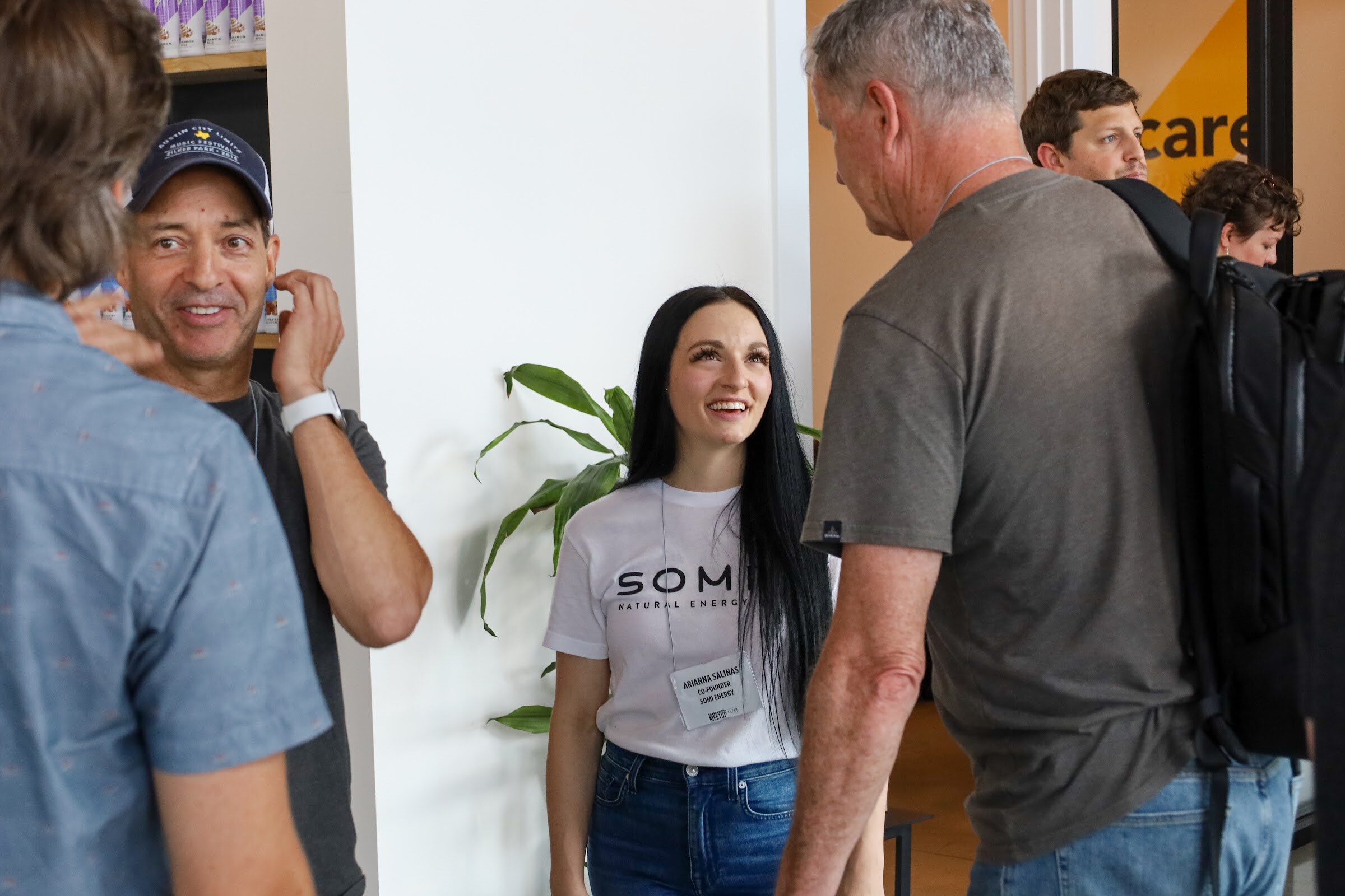 A smiling woman with long black hair in a white Somi Natural Energy t-shirt with a name badge looks toward arriving guests, including a man in a navy cap and a man in a gray t-shirt with a black backpack, with a plant and a yellow wall behind her.